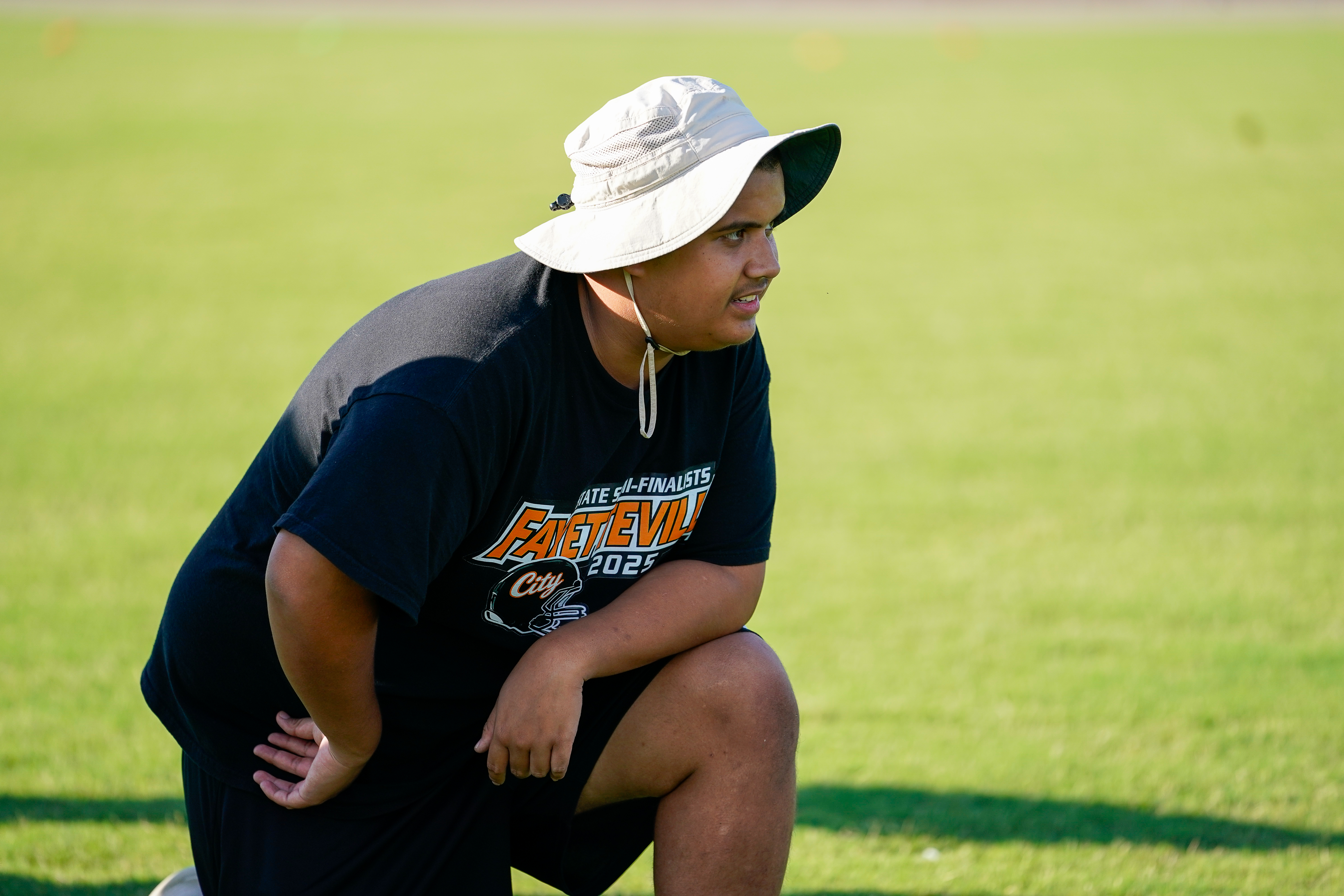 A young person in a black t-shirt and bucket hat kneels on a green field, looking off to the side with a blurred grassy background.