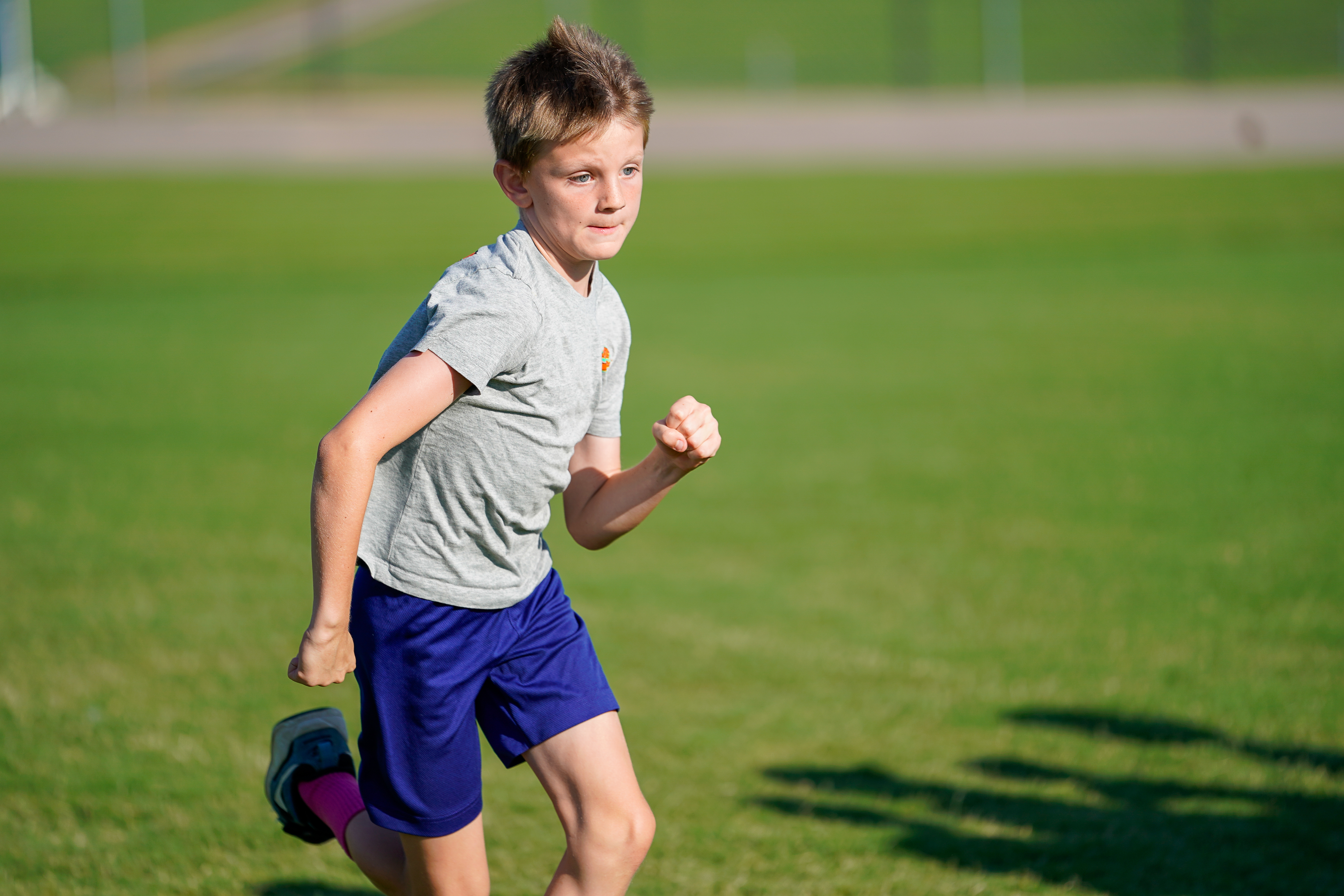A young boy in a grey t-shirt and blue shorts runs across a grassy field with a blurred green background.