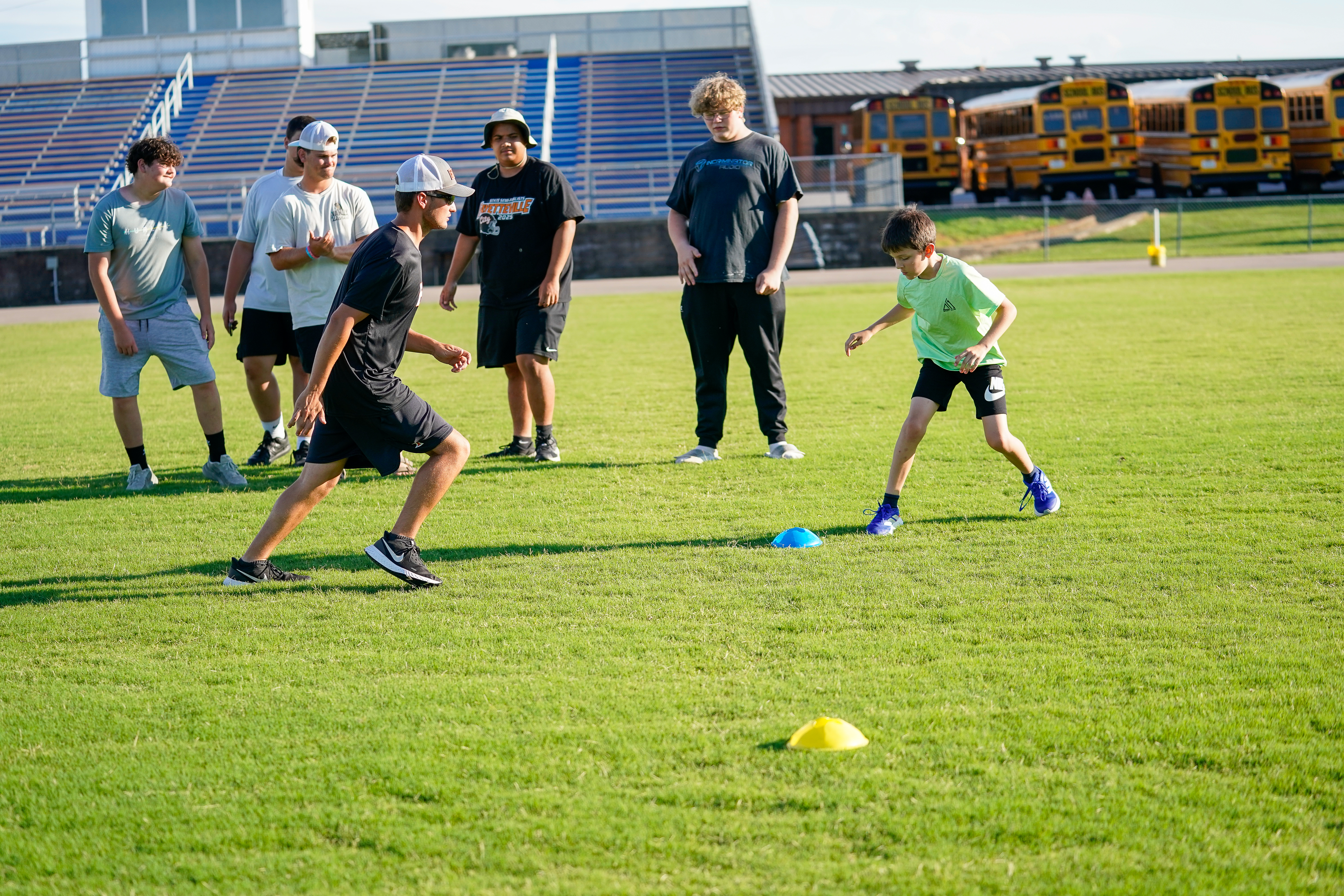 Two boys practice drills on a grassy field with cones, while a group of older boys watches and yellow school buses are parked in the background.