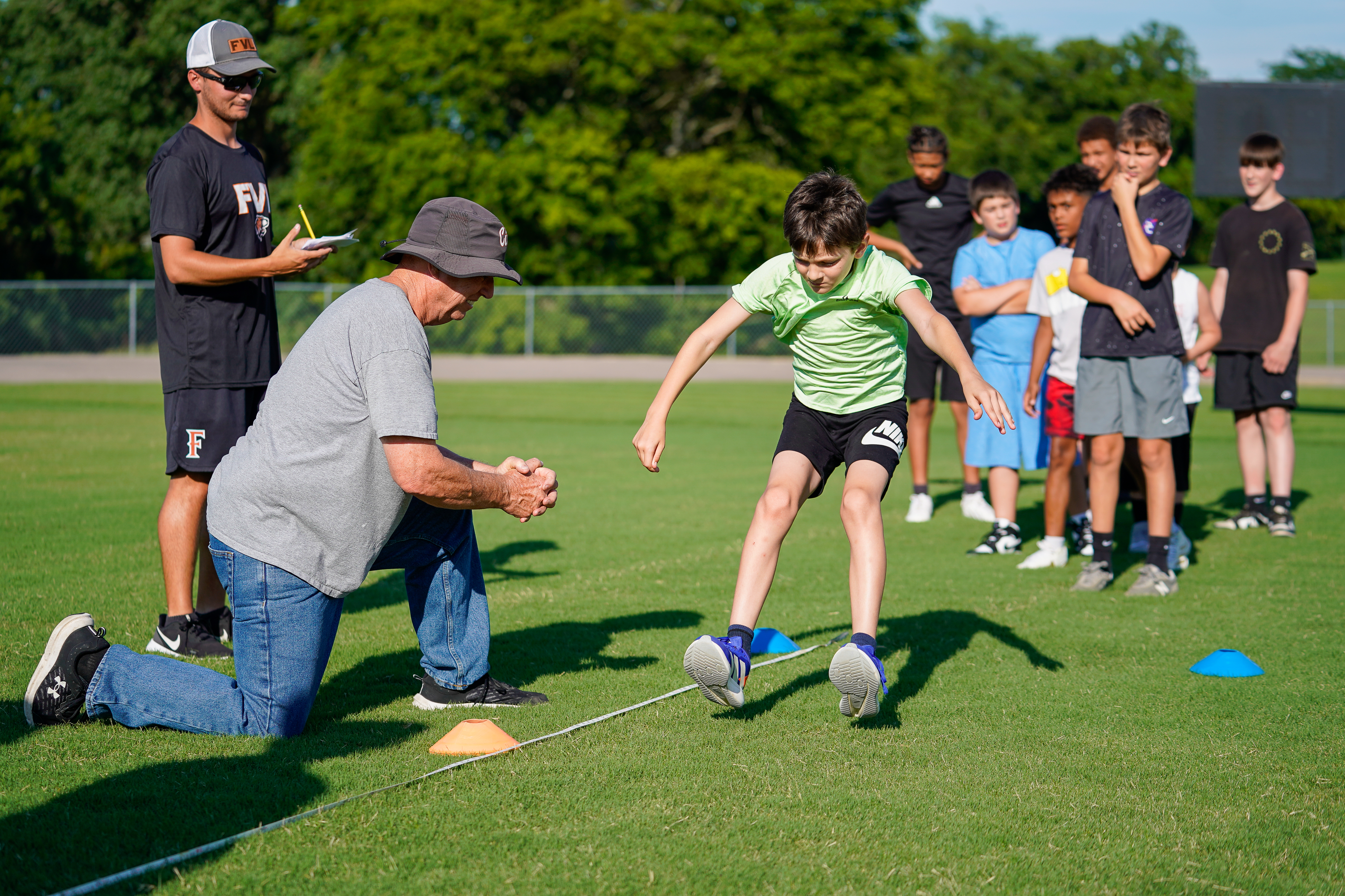 A young boy in a green shirt jumps over a line on a grassy field while an older man coaches him, with a group of other boys and a coach standing in the background under a tree-lined sky.