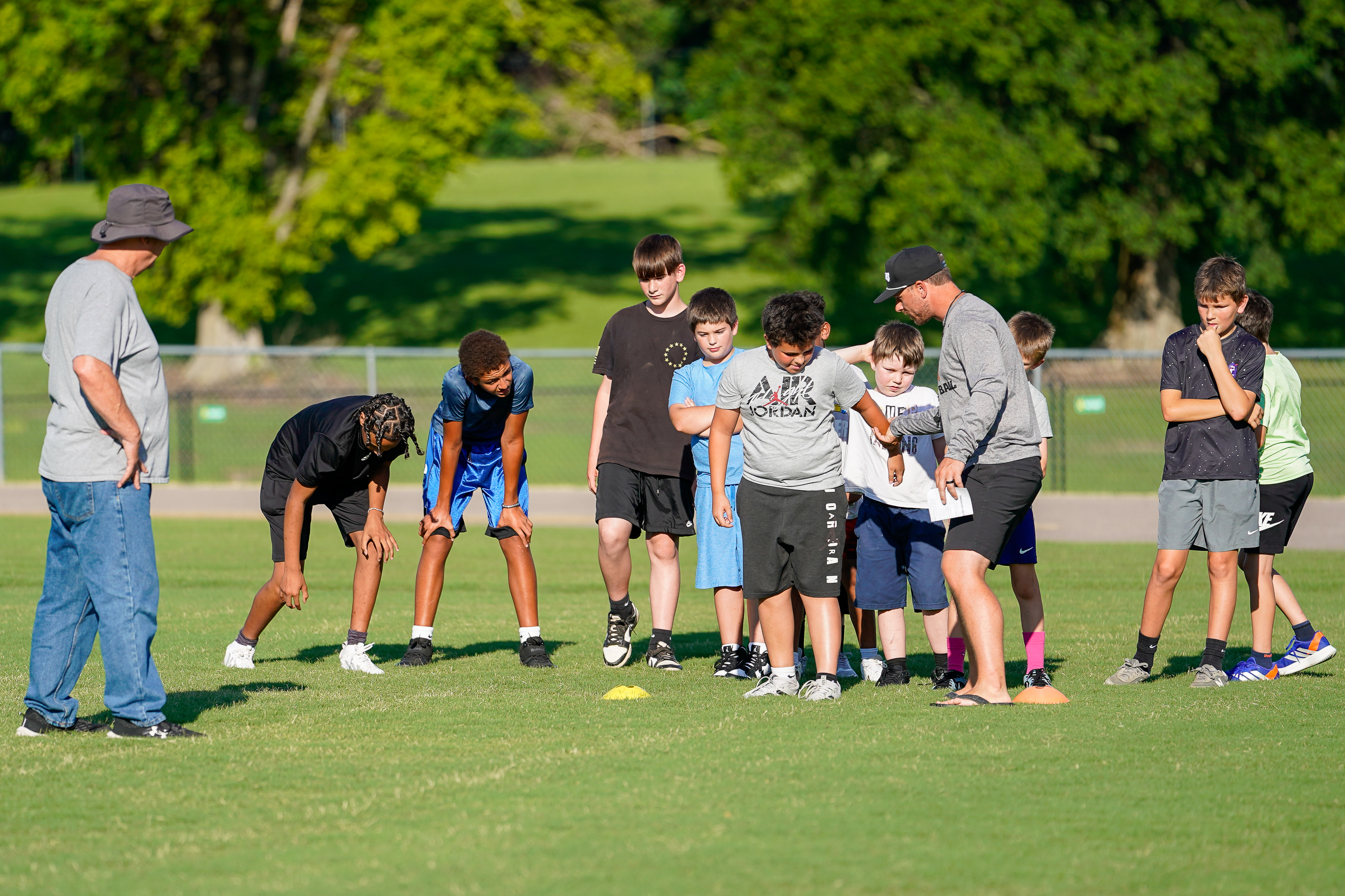 A group of young boys practice football drills on a grassy field with trees in the background.