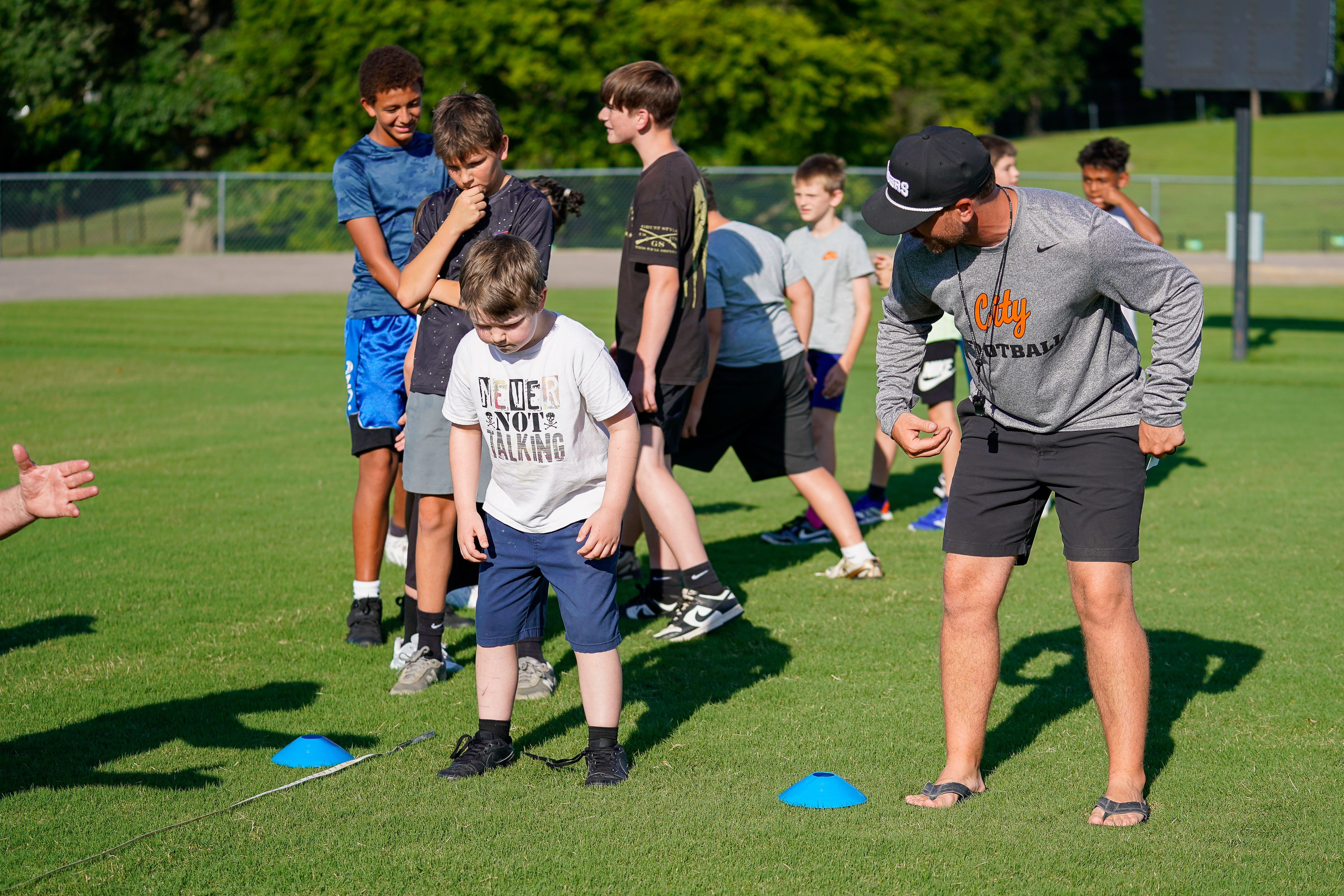 A group of young boys and a coach are on a grassy field with blue cones in the foreground, while trees and a fence are visible in the background.