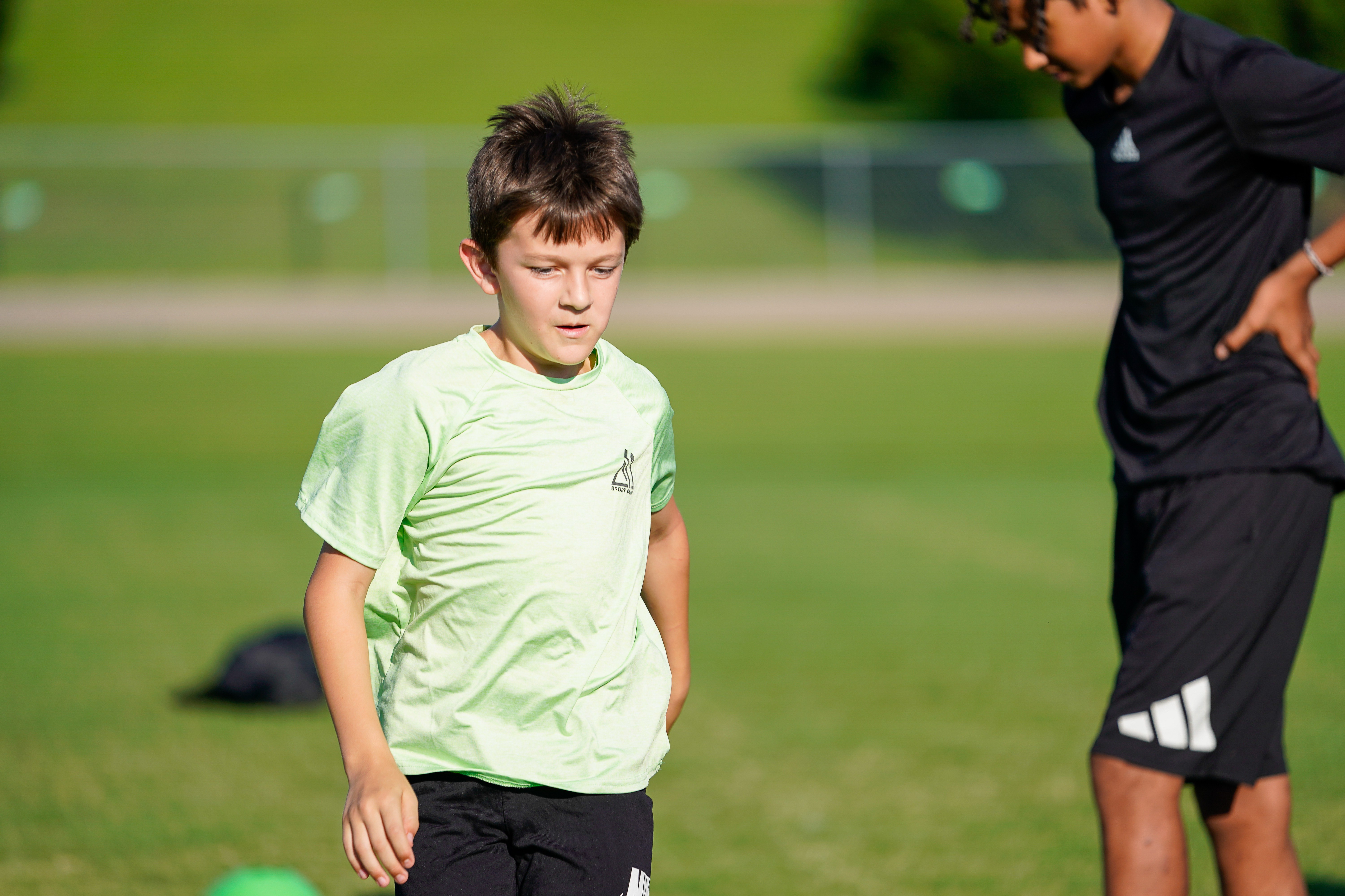 A young boy in a light green shirt and black shorts stands on a grassy field in the foreground, while another boy in a black shirt and shorts is partially visible to his right, with a blurred green background suggesting a sports field.