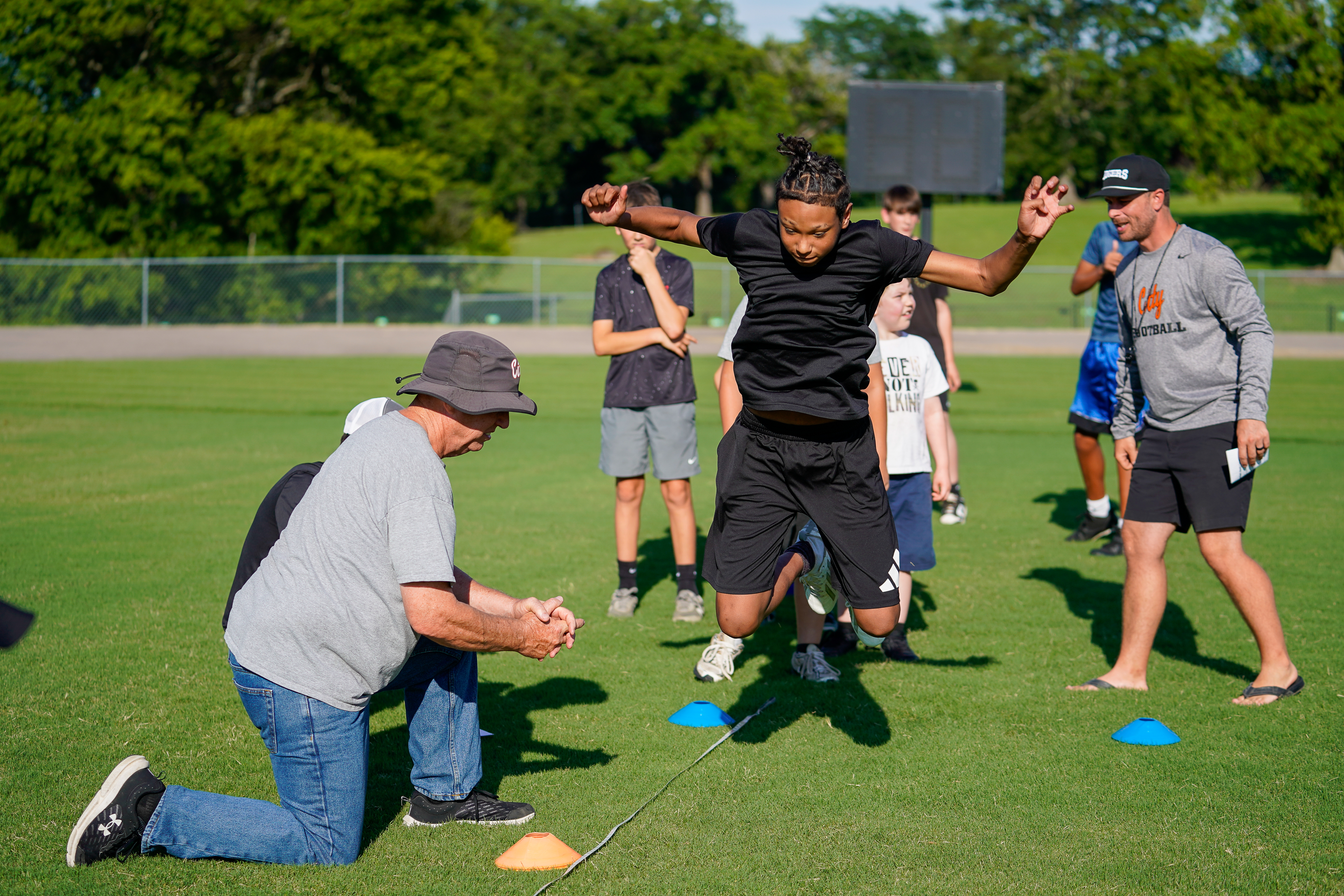 A young man in mid-air jumps over a rope on a grassy field, with coaches and other athletes observing in the background.