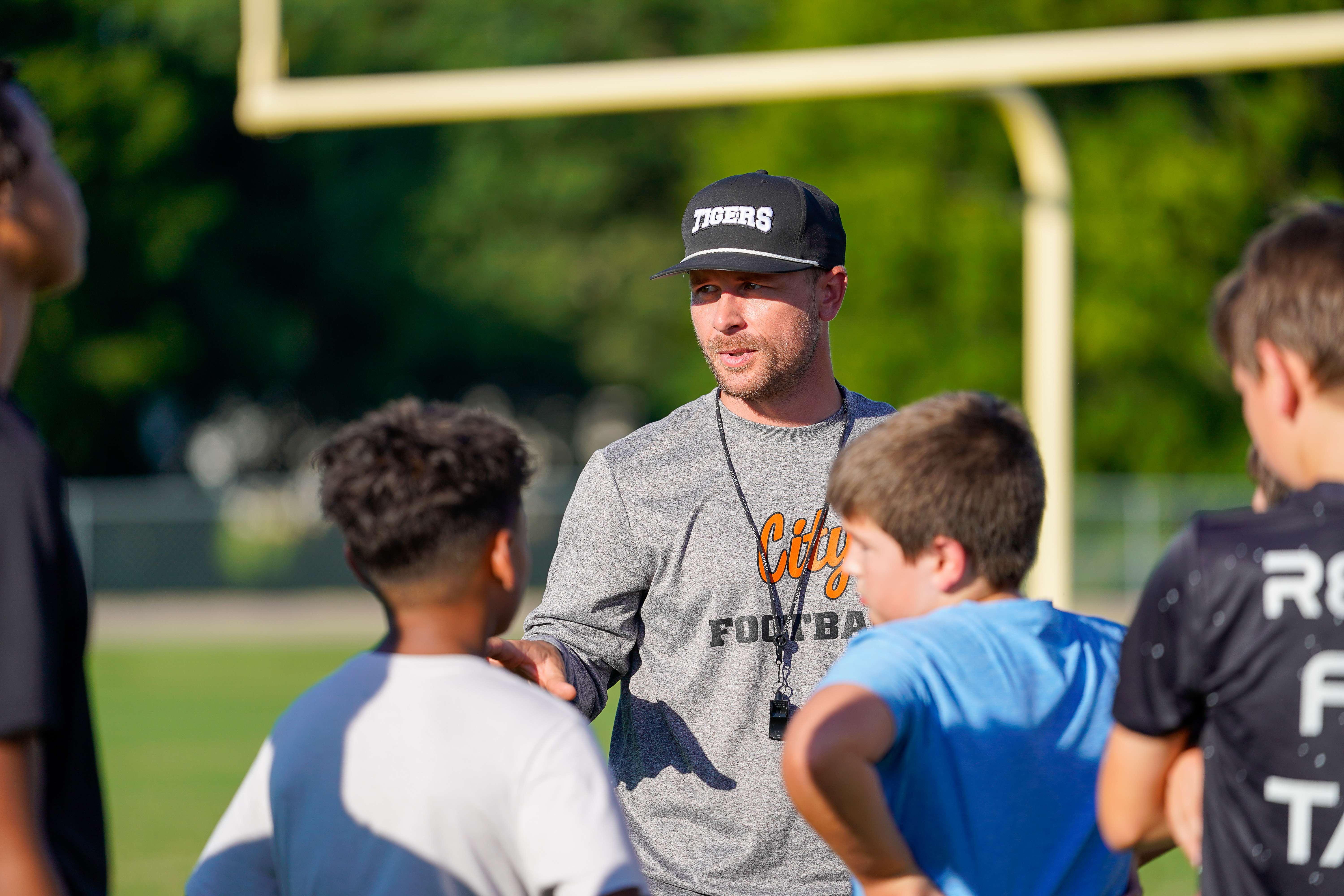 A football coach wearing a "Tigers" hat and a gray "City Football" shirt talks to a group of young players on a field with a goalpost in the background.