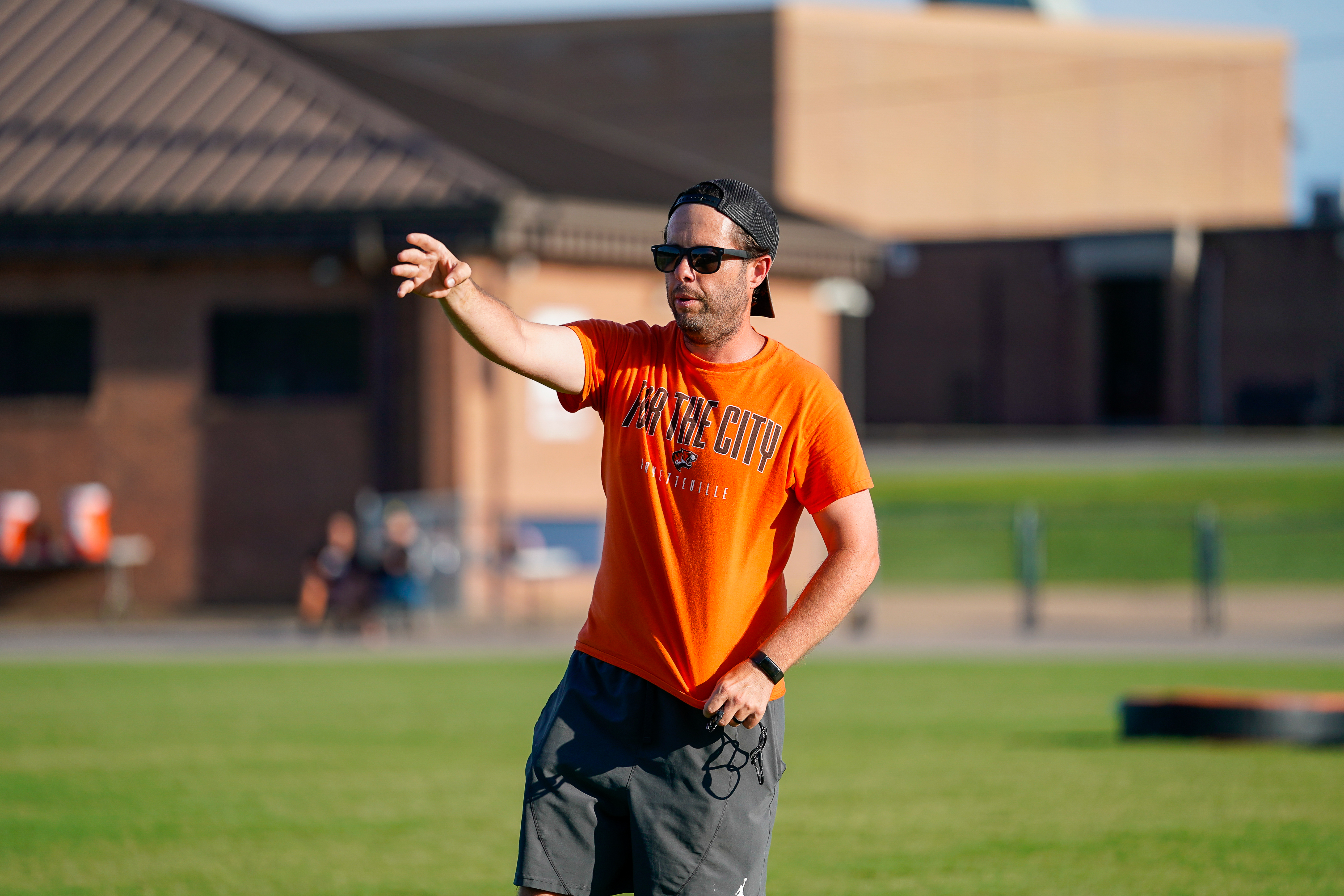 A man in an orange t-shirt and sunglasses gestures on a grassy field with buildings in the background.