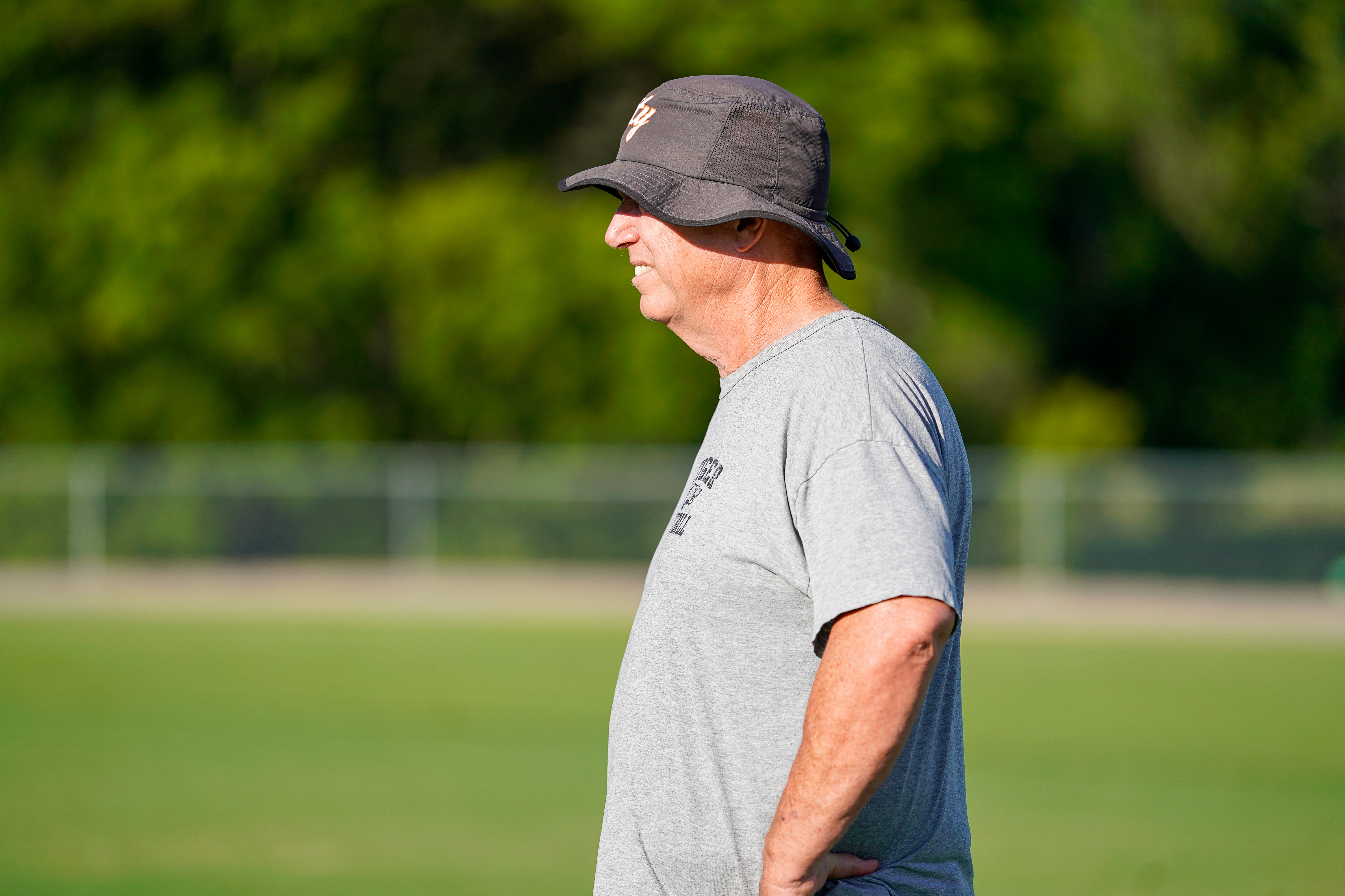 A man in a bucket hat and t-shirt stands on a grassy field with a blurred treeline in the background.