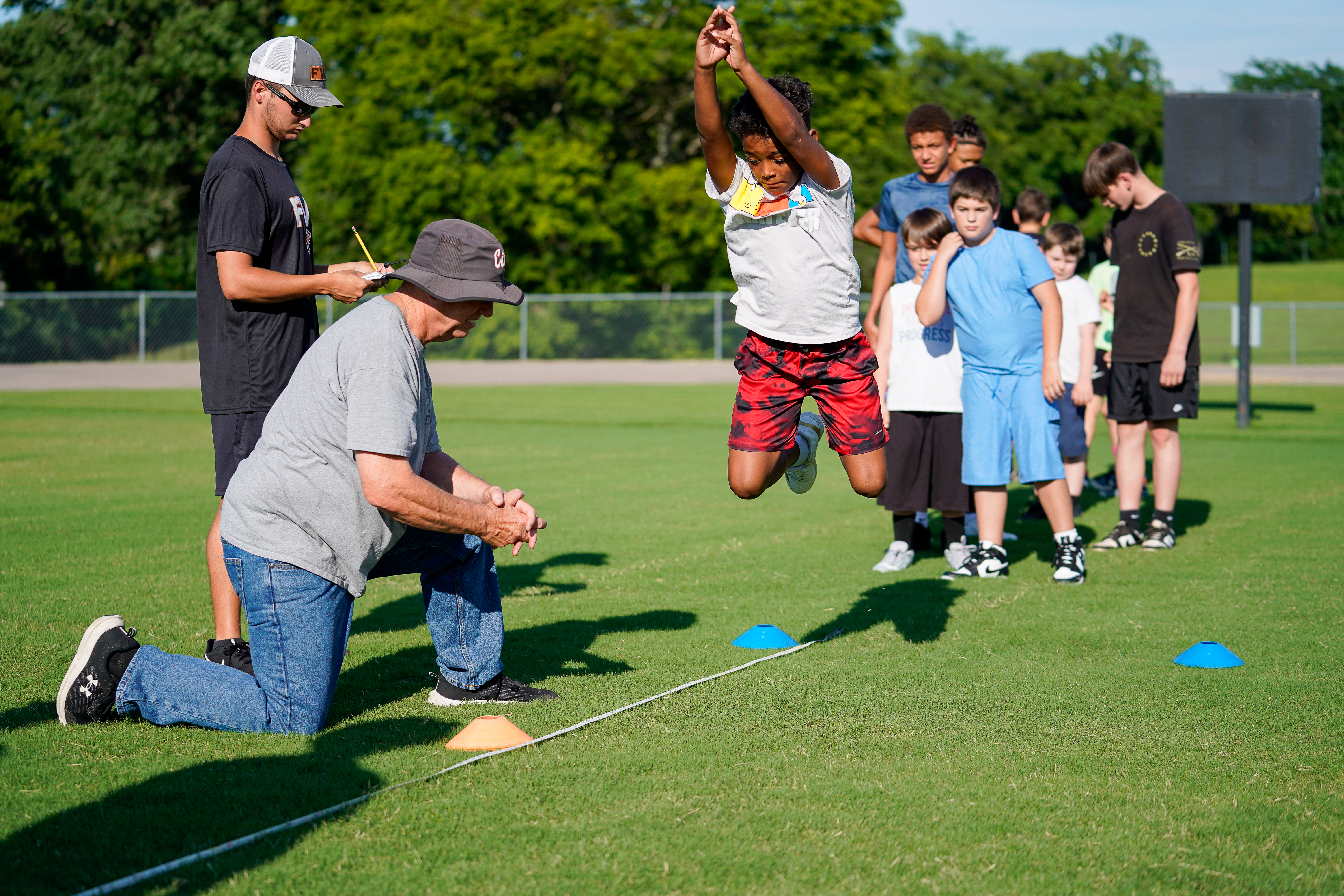 A young boy leaps in the air during a track and field event on a grassy field, with coaches and other children watching in the background.