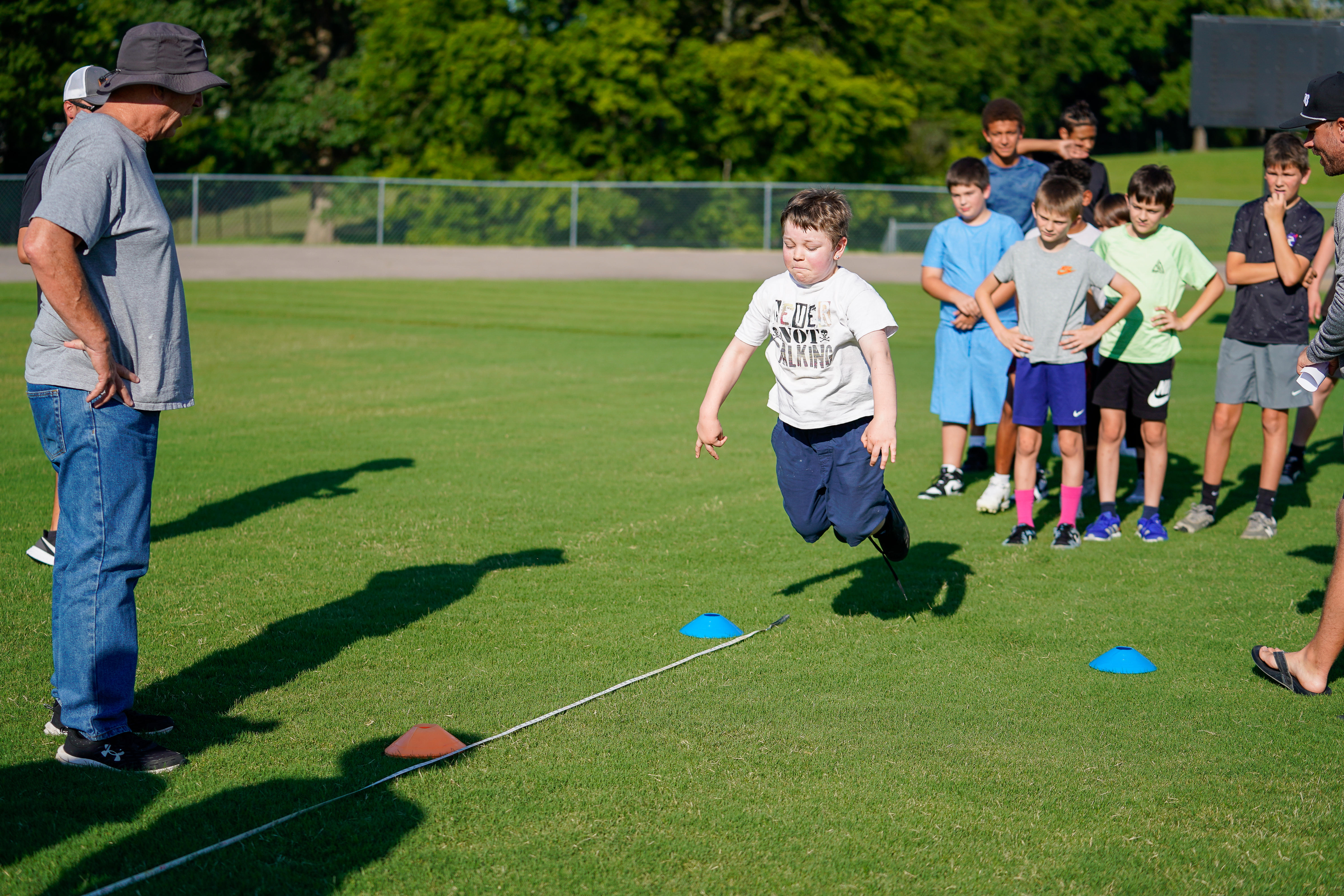 A young boy jumps over cones on a grassy field while an adult watches and other children stand in the background.