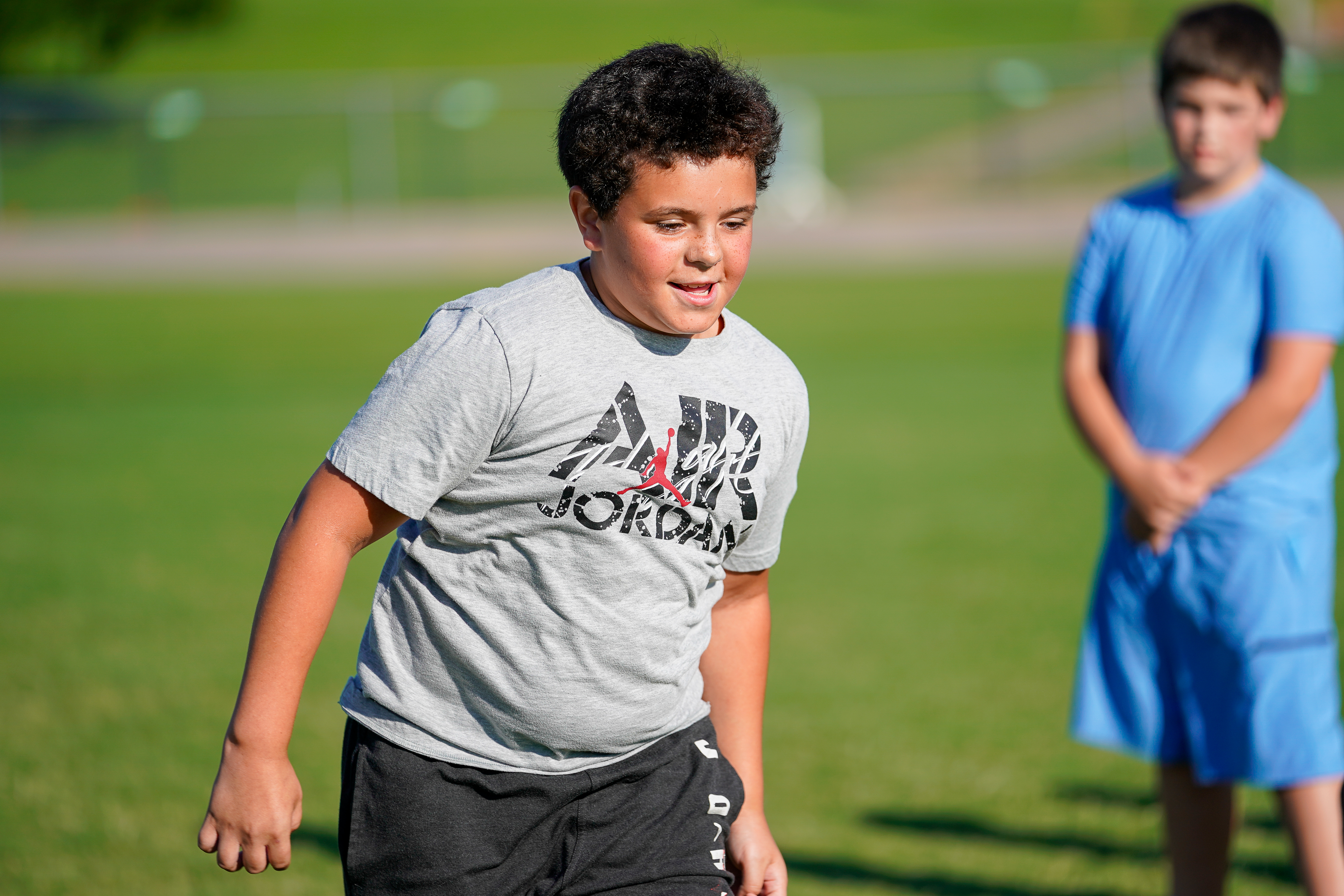 A young boy in a grey t-shirt and black shorts is in the foreground, while another boy in a blue t-shirt and shorts stands in the blurred green background.