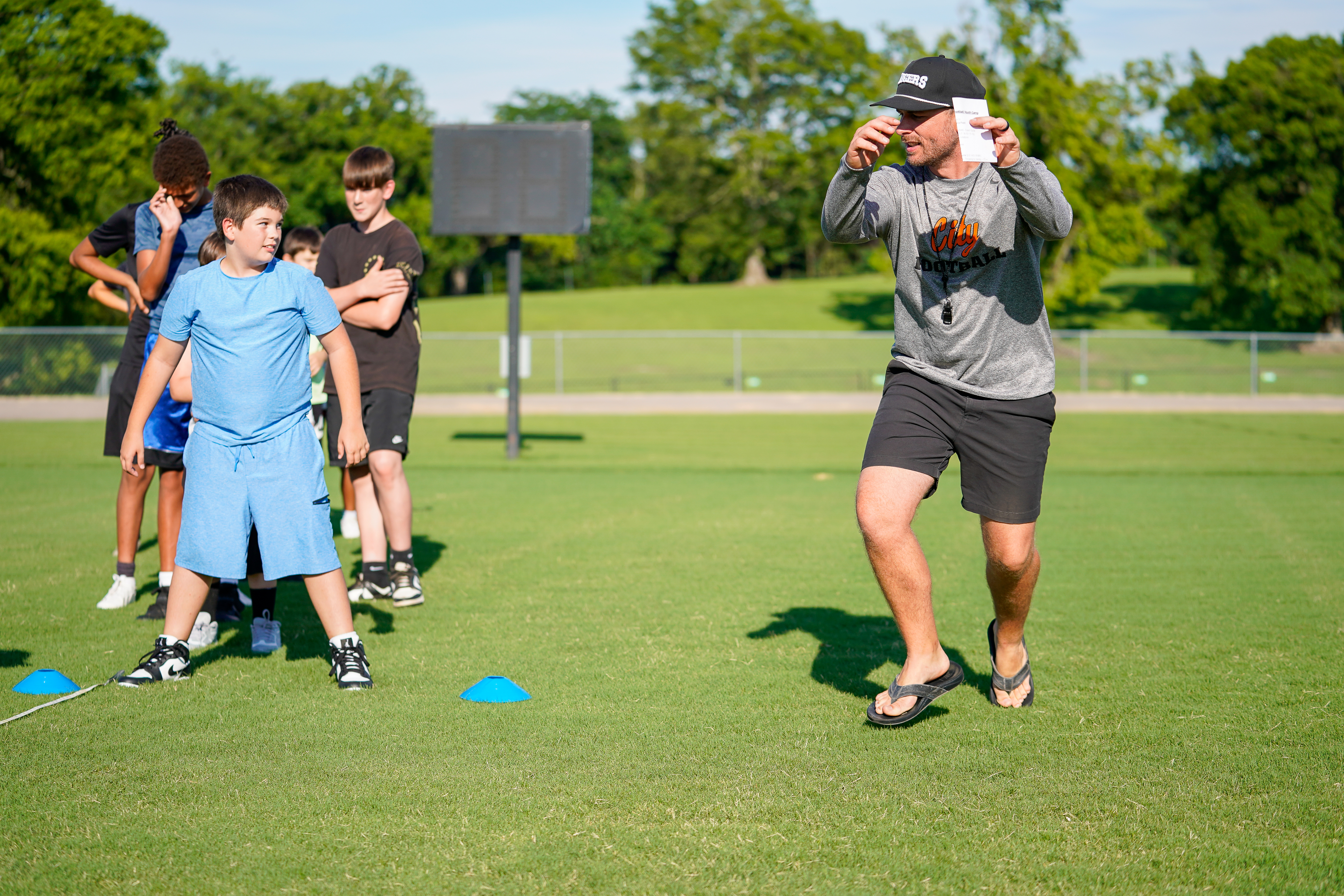 A coach holds up a card to a group of young athletes on a grassy field with trees in the background.
