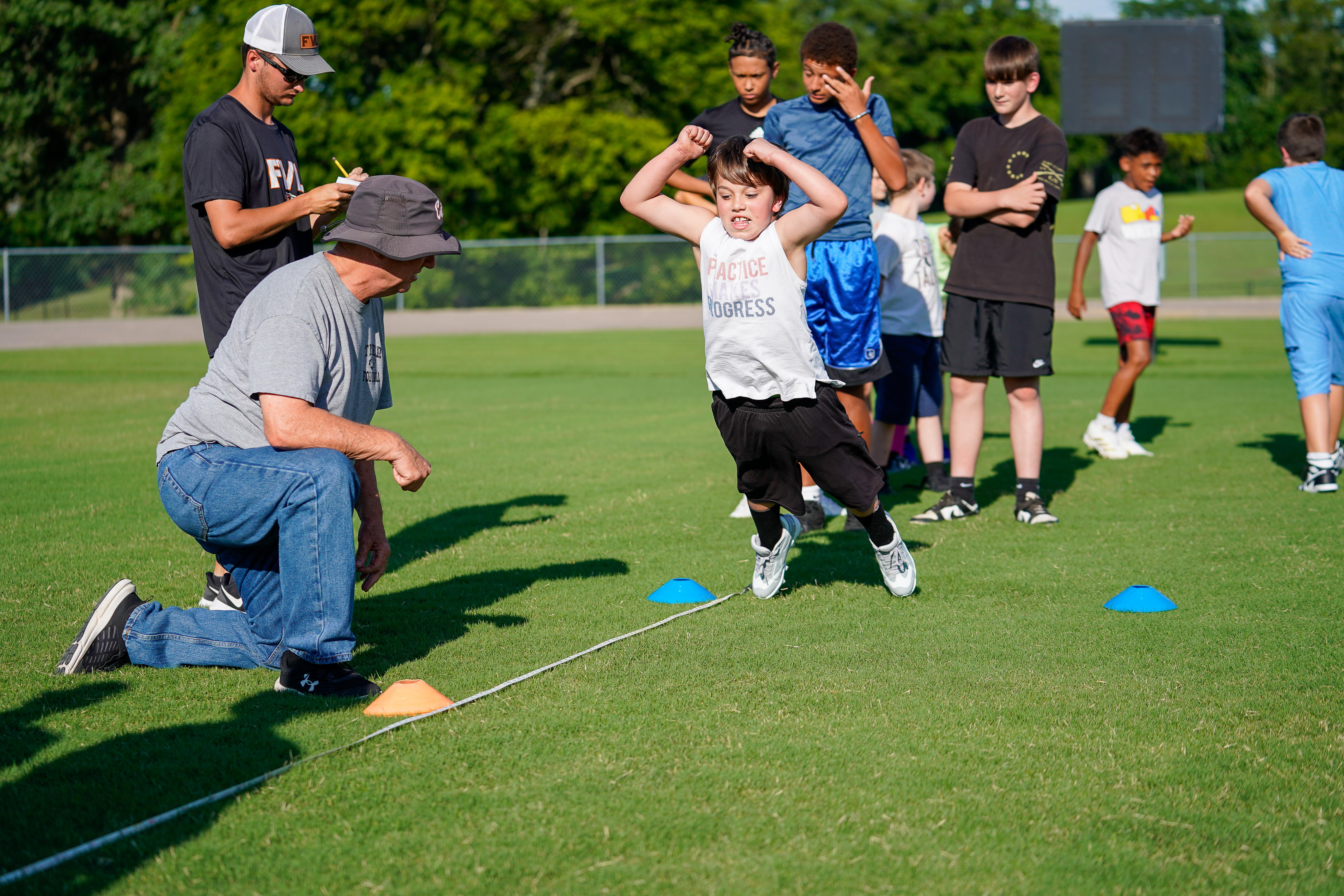 A young boy jumps over cones on a grassy field while an older man watches, with other children and a coach in the background.