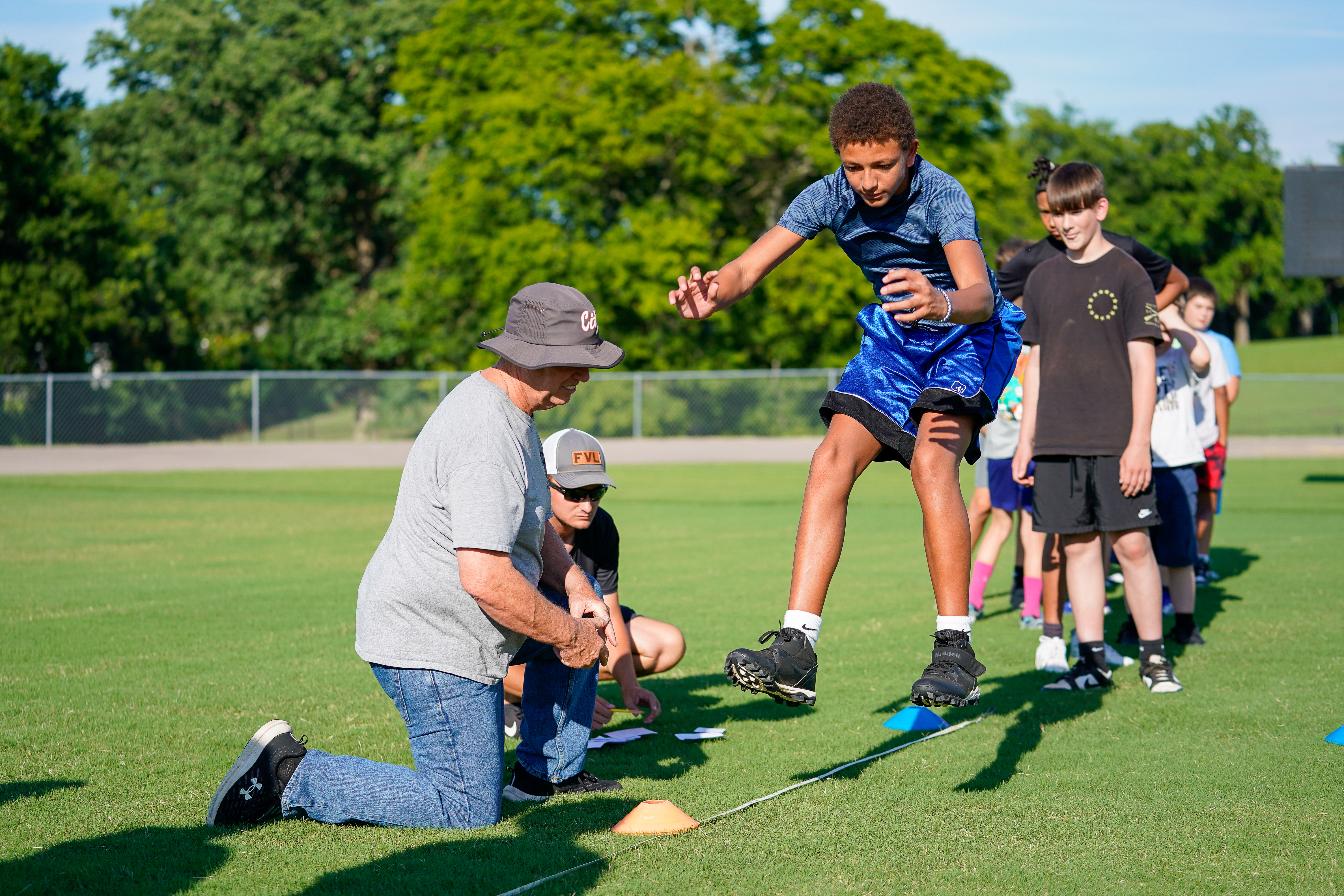 A coach instructs young athletes on a grassy field with trees in the background.