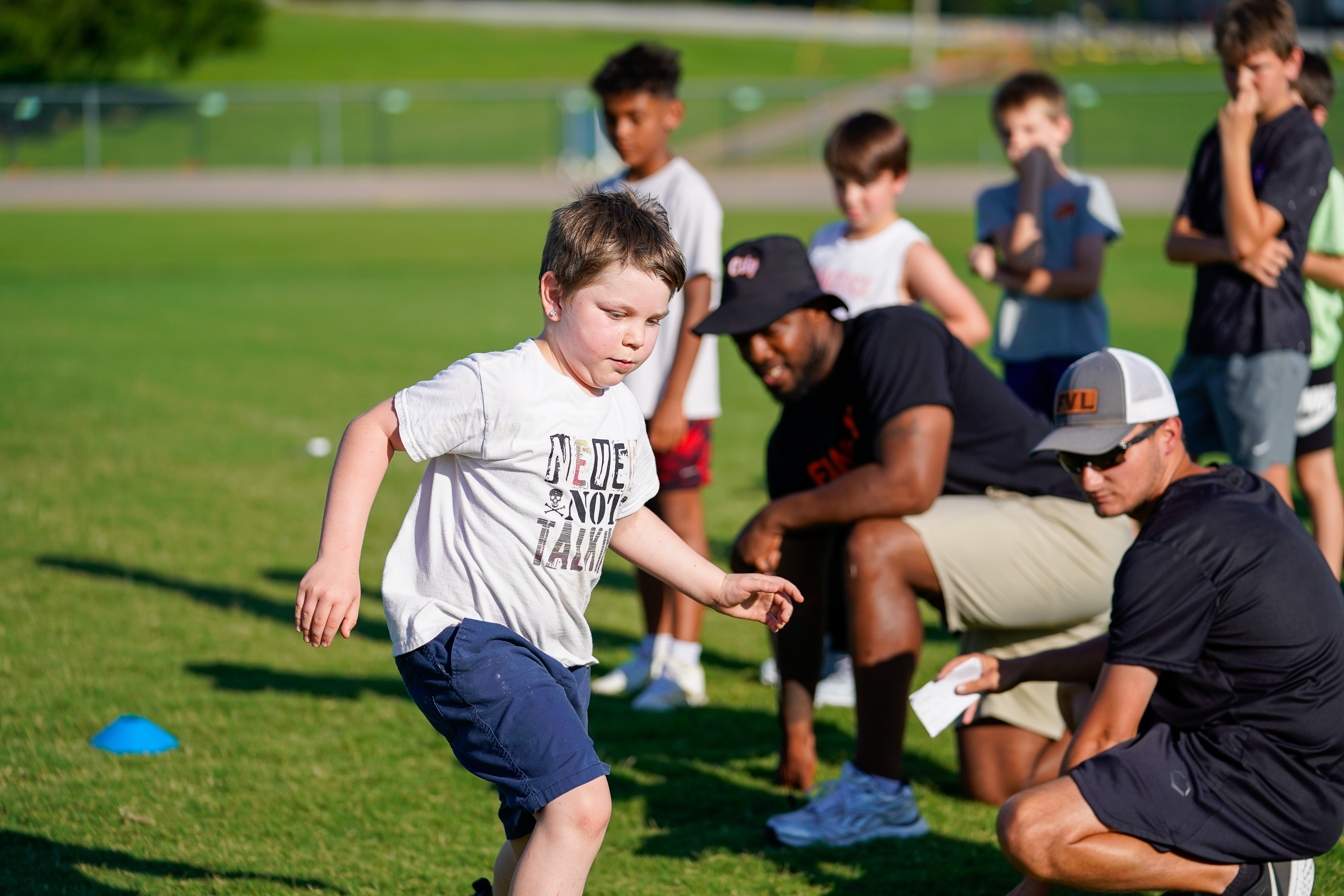 A young boy in the foreground runs on a grassy field while coaches and other children watch in the background.