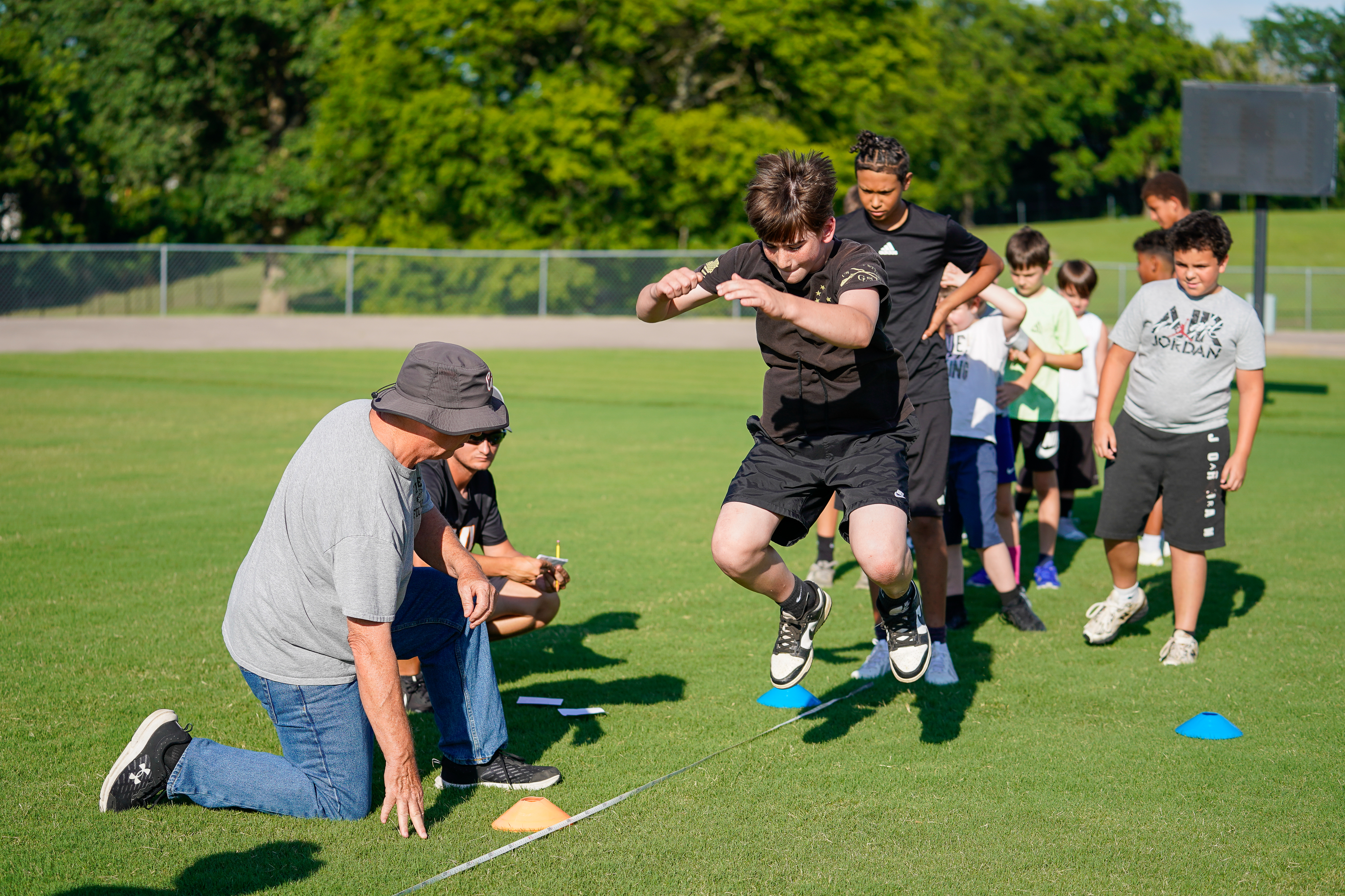 A young athlete jumps over cones on a grassy field while a coach watches, with other children and trees in the background.