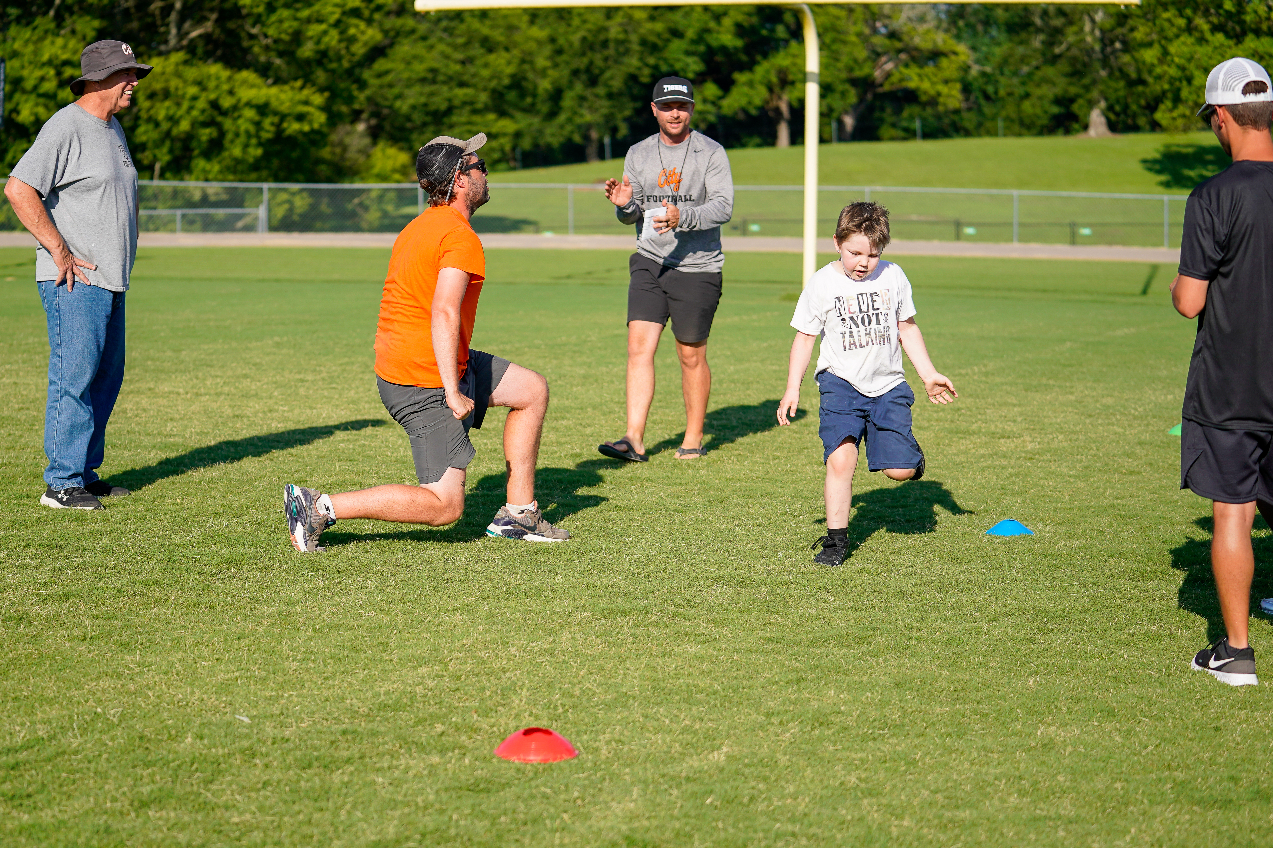 A young boy runs on a grassy field with coaches and spectators in the background.