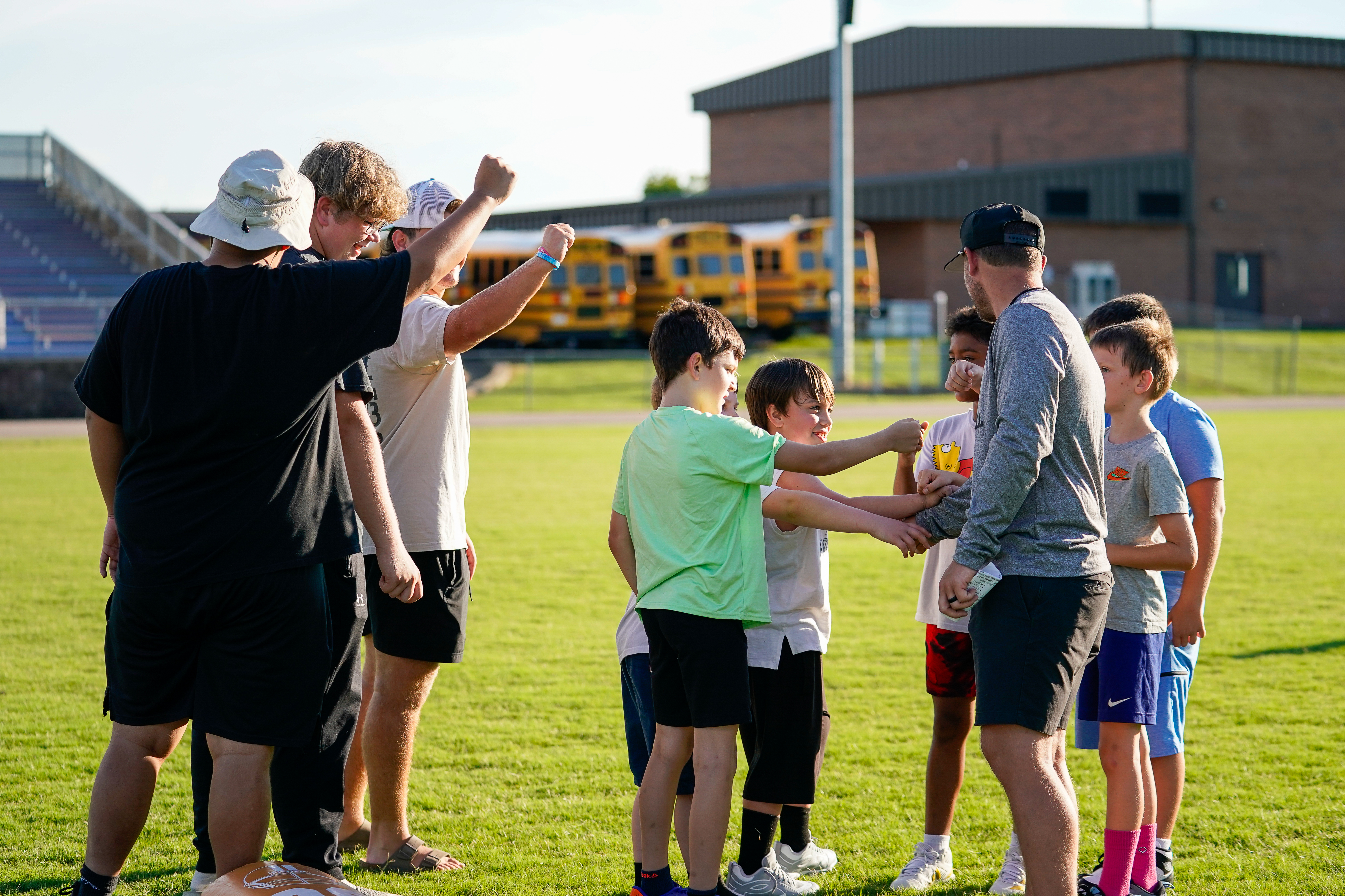 A group of boys on a grassy field, with a line of yellow school buses and a stadium in the background.
