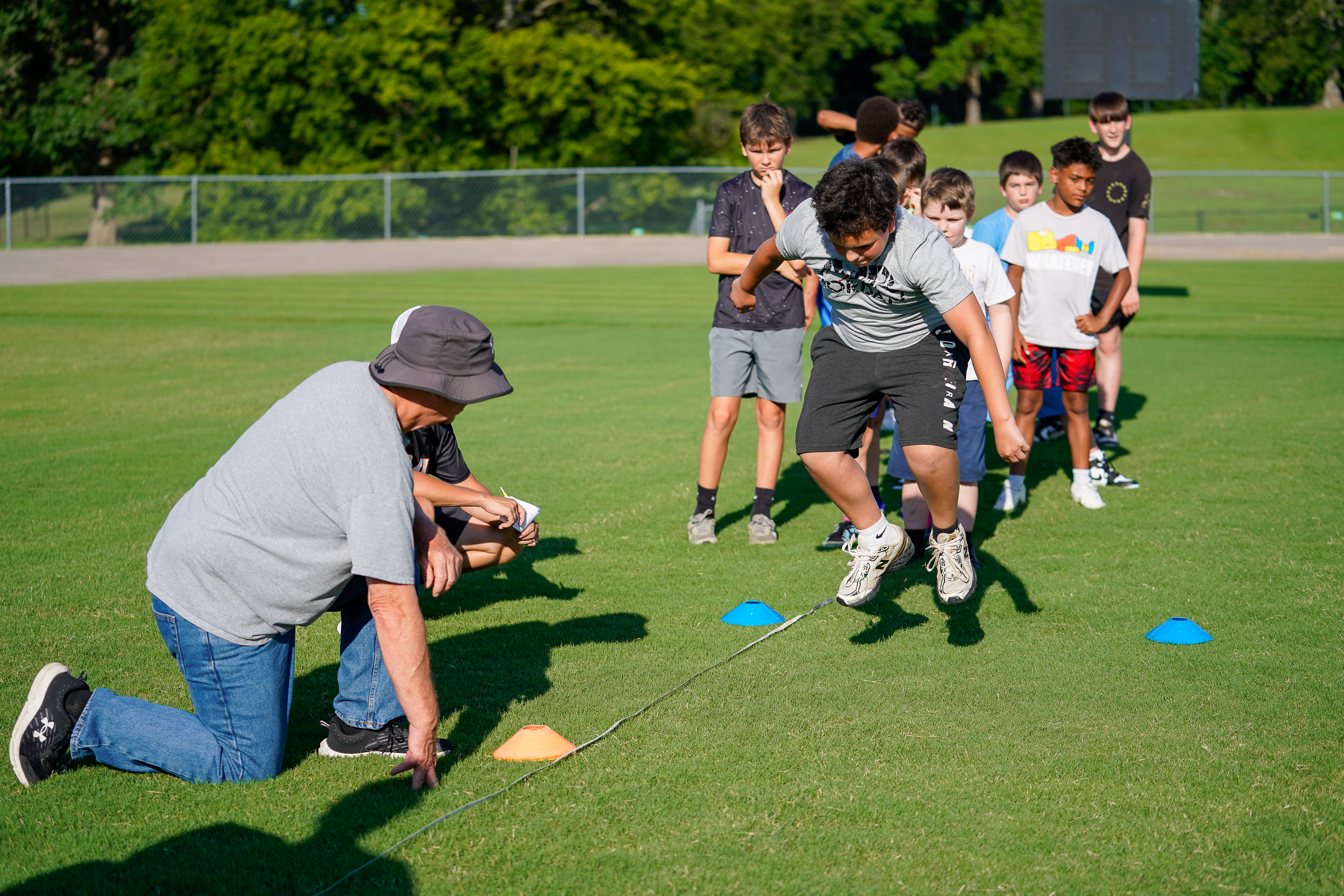 A man in a hat coaches a group of boys performing agility drills on a grassy field with trees and a fence in the background.