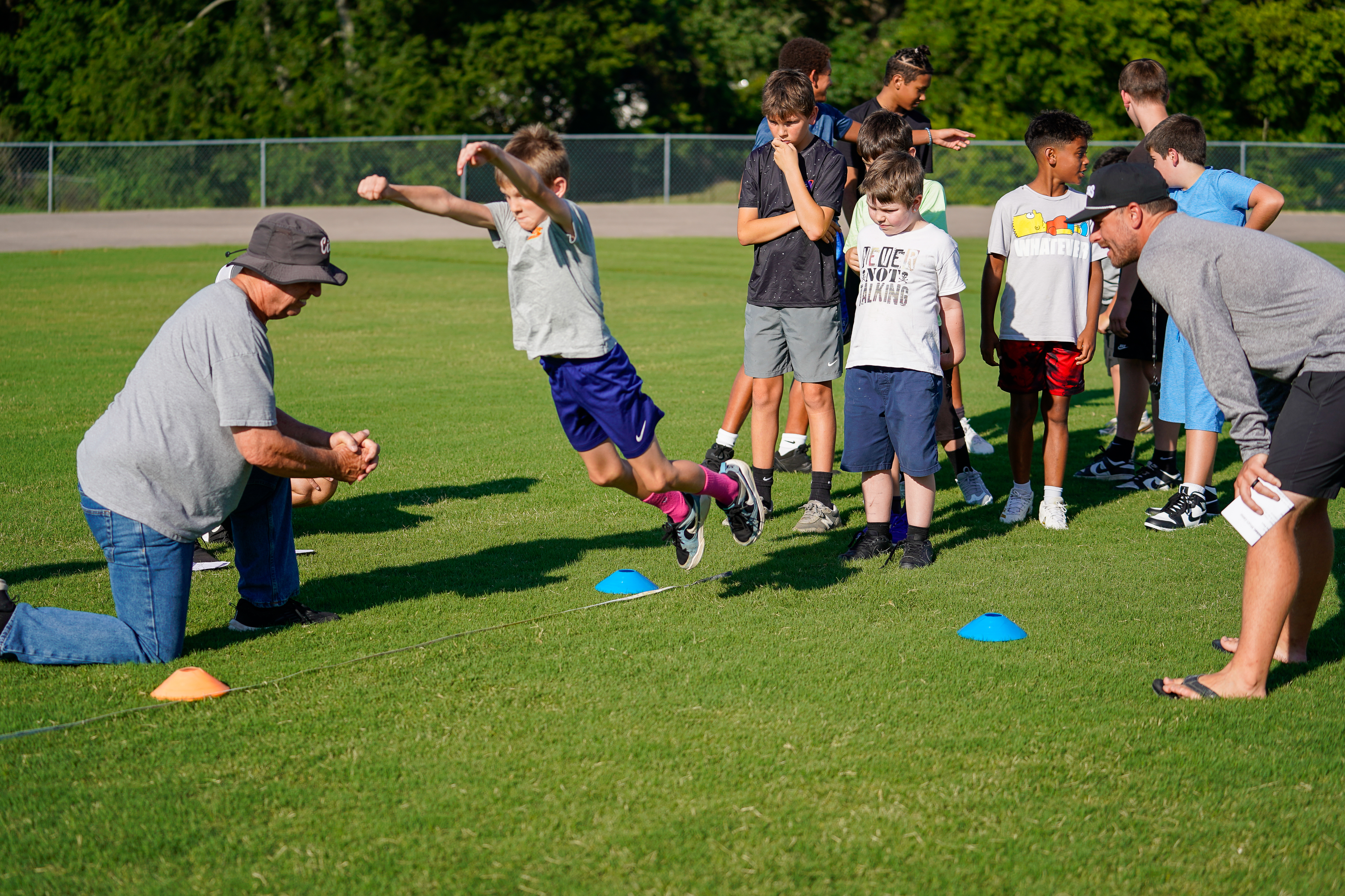A young boy leaps over cones on a grassy field while coaches and other boys watch in the background.
