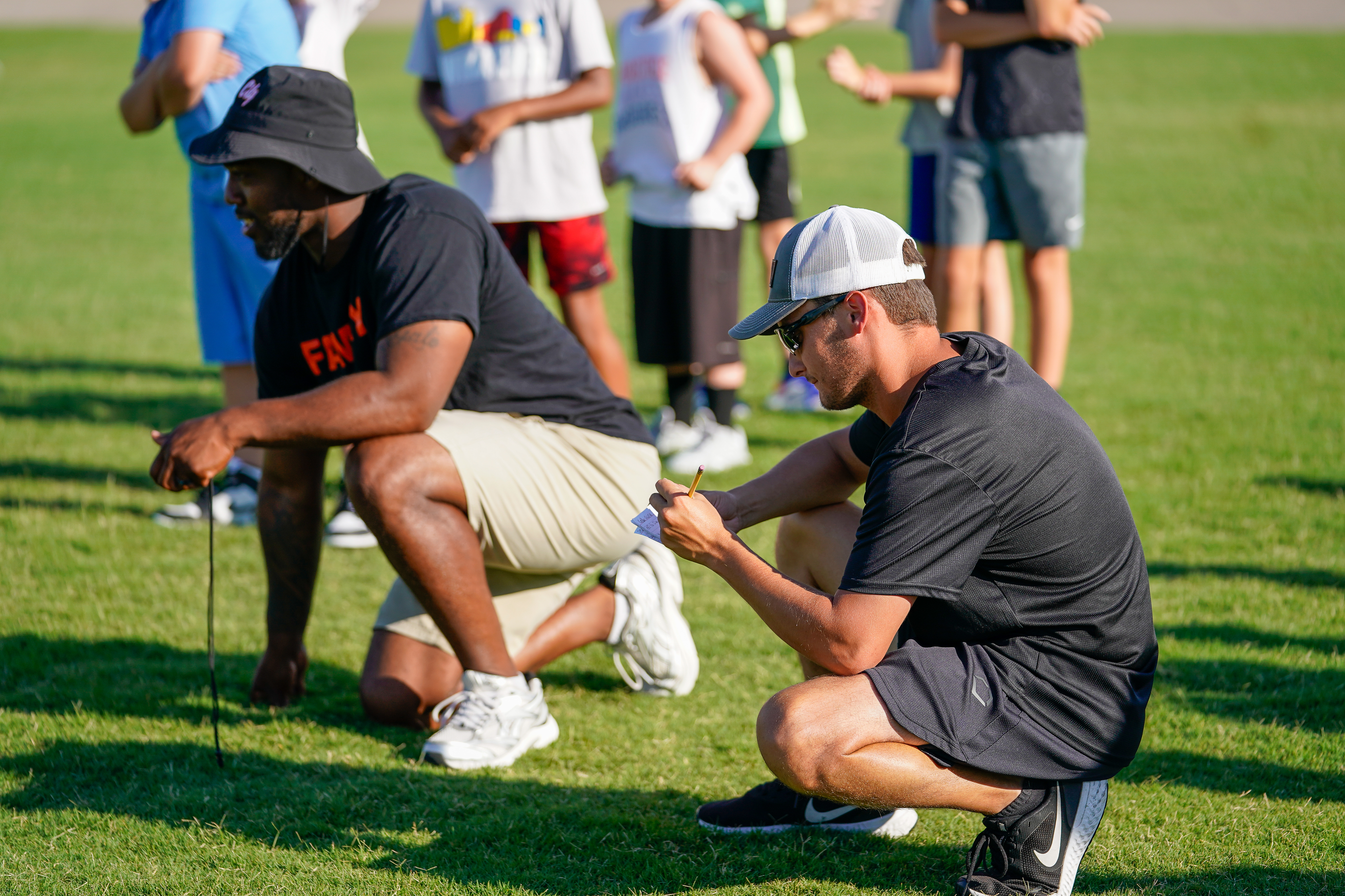 Two coaches in the foreground crouch on a grassy field, observing a group of children in the blurred background.
