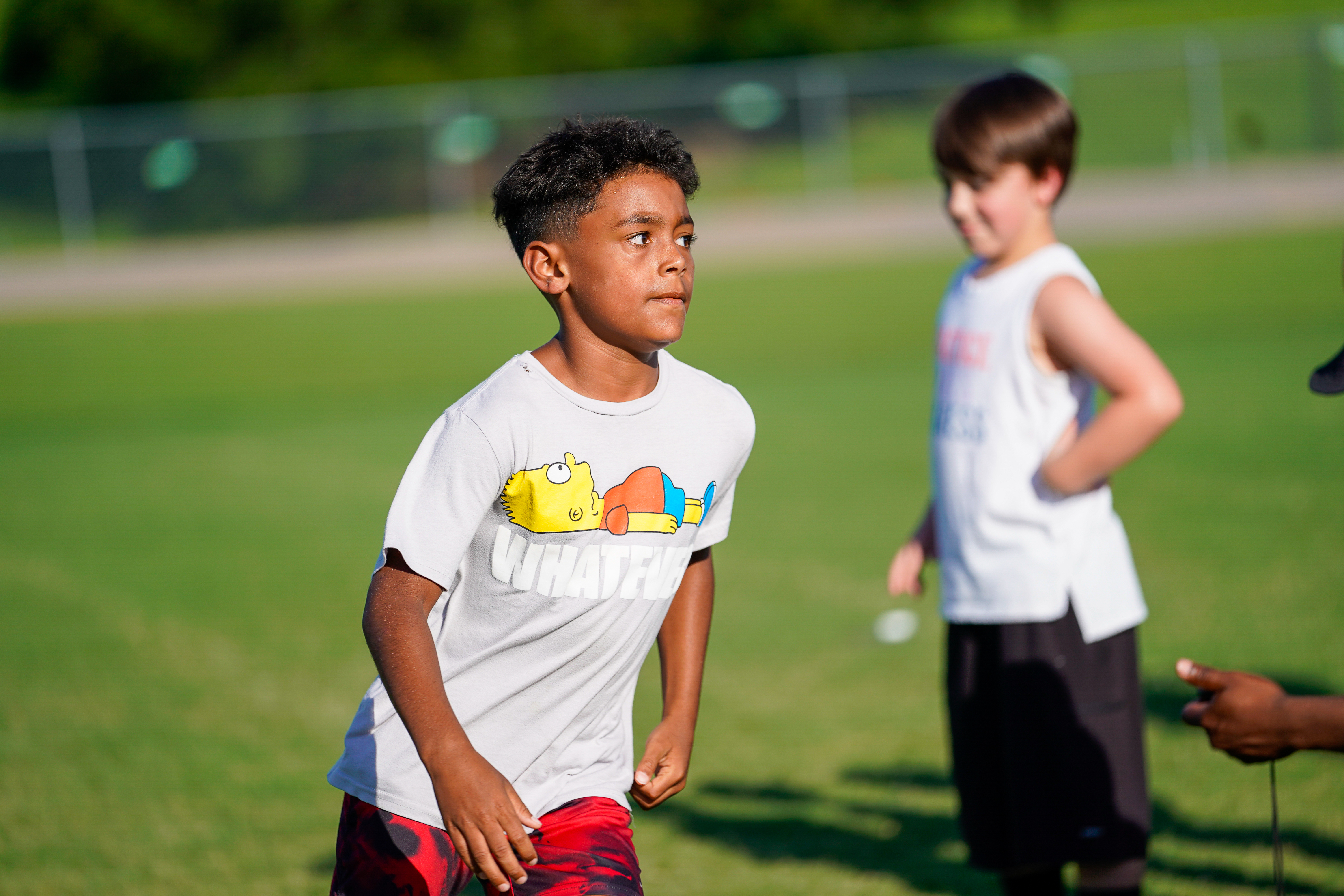 A young boy in a gray t-shirt and red shorts stands on a grassy field with another boy blurred in the background.
