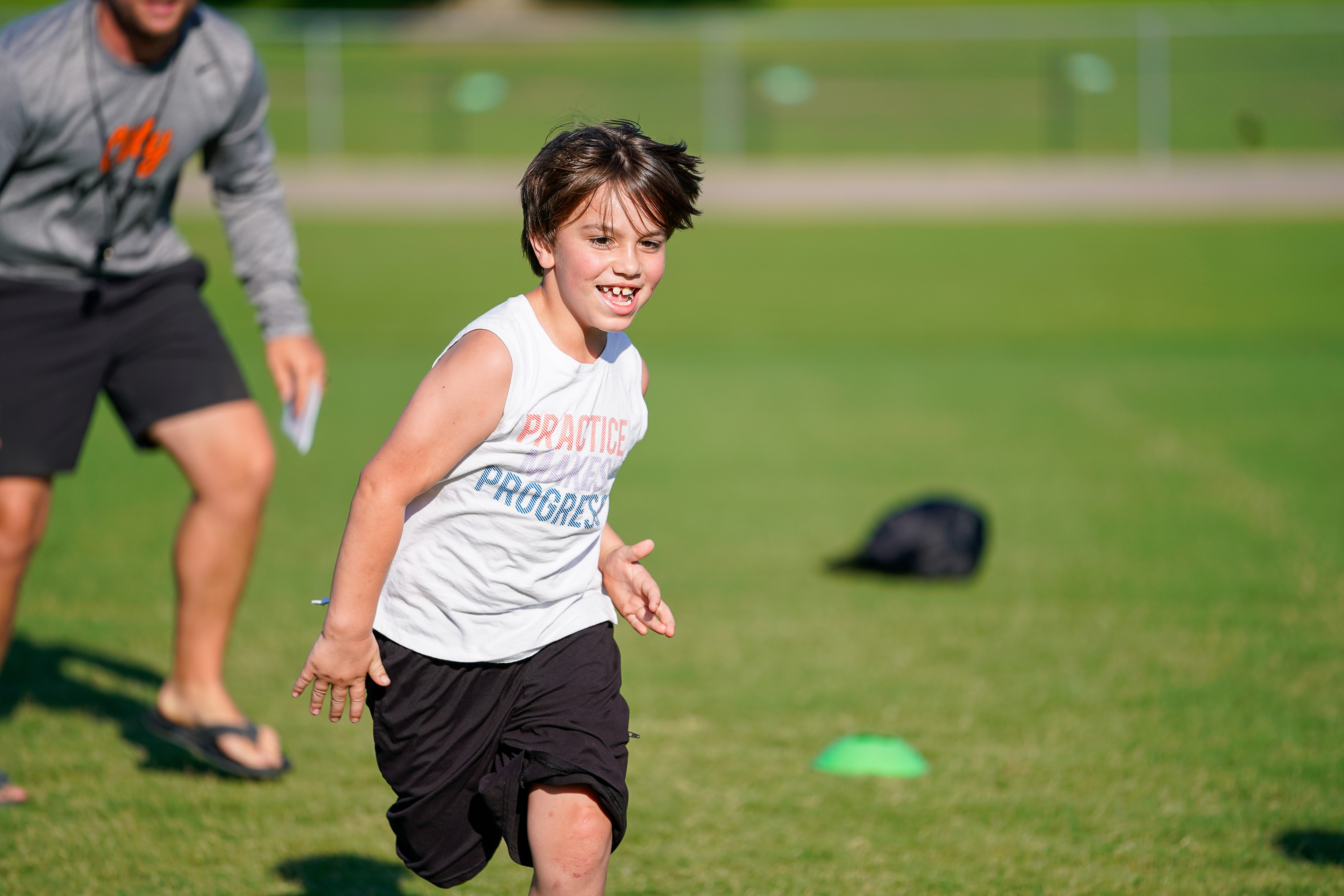 A young boy in a white tank top and black shorts runs on a grassy field with a blurred figure in athletic wear behind him and a green cone in the background.