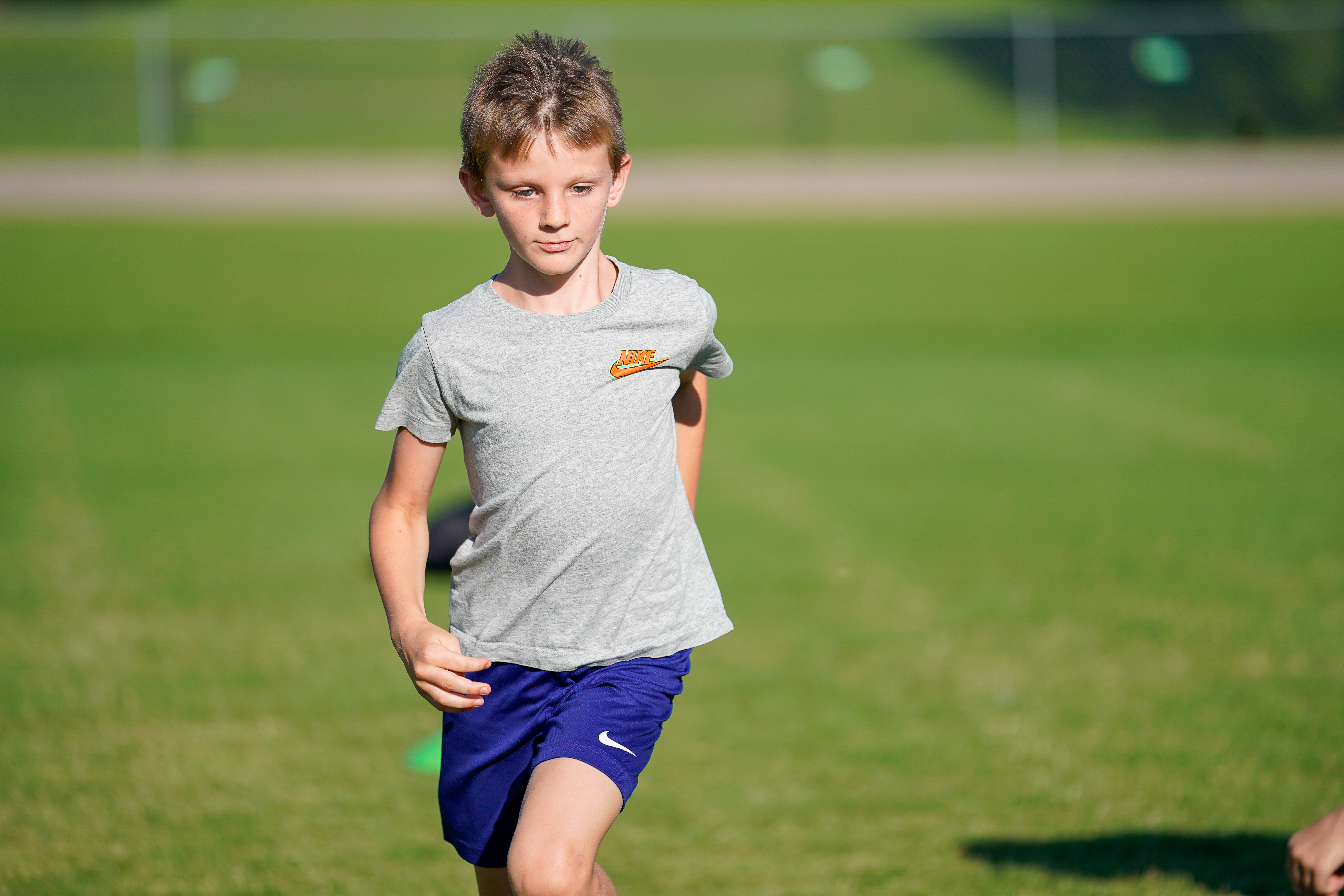 A young boy in a gray t-shirt and blue shorts runs on a grassy field with a blurred green background.