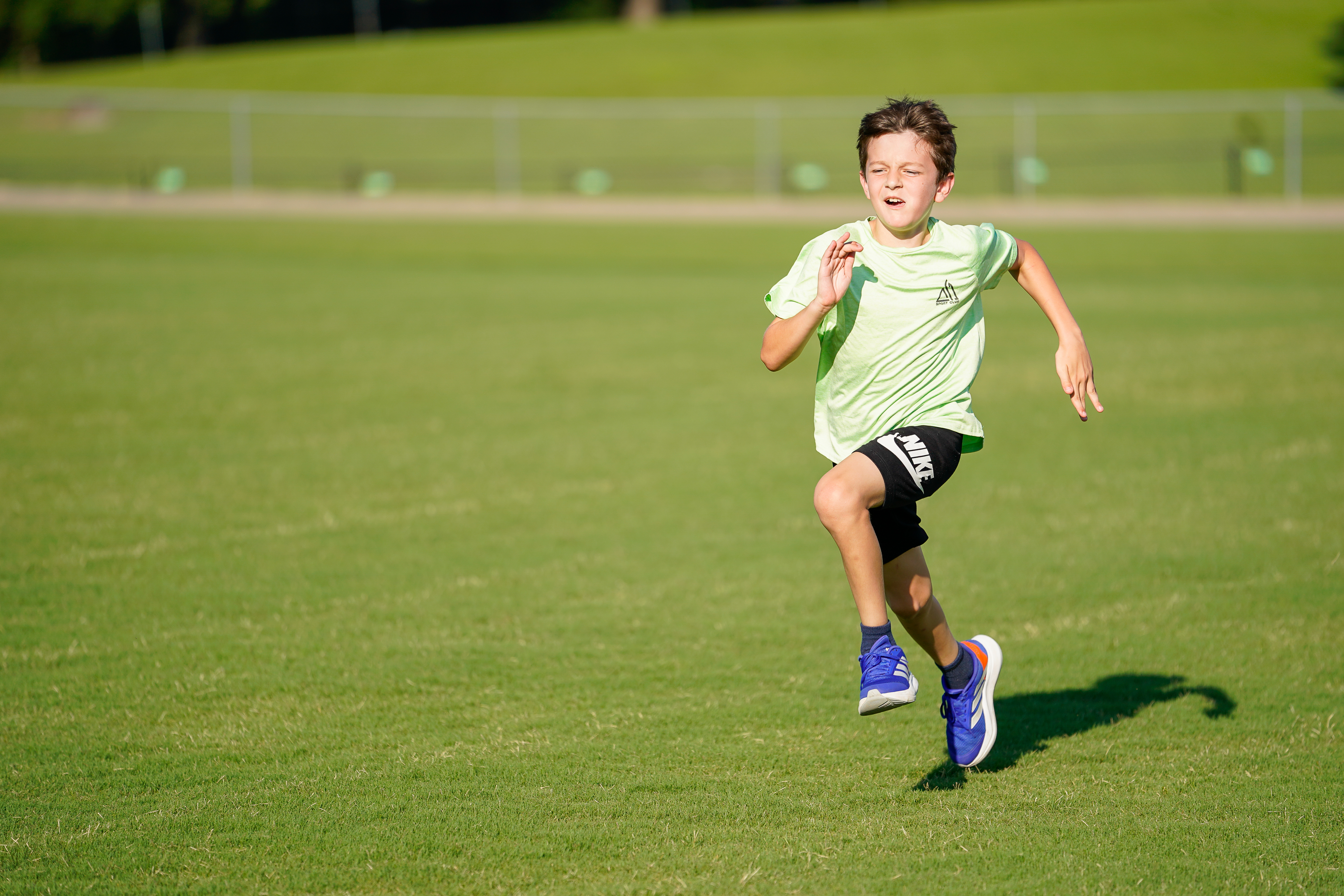 A young boy in athletic wear sprints across a grassy field with a blurred fence and trees in the background.