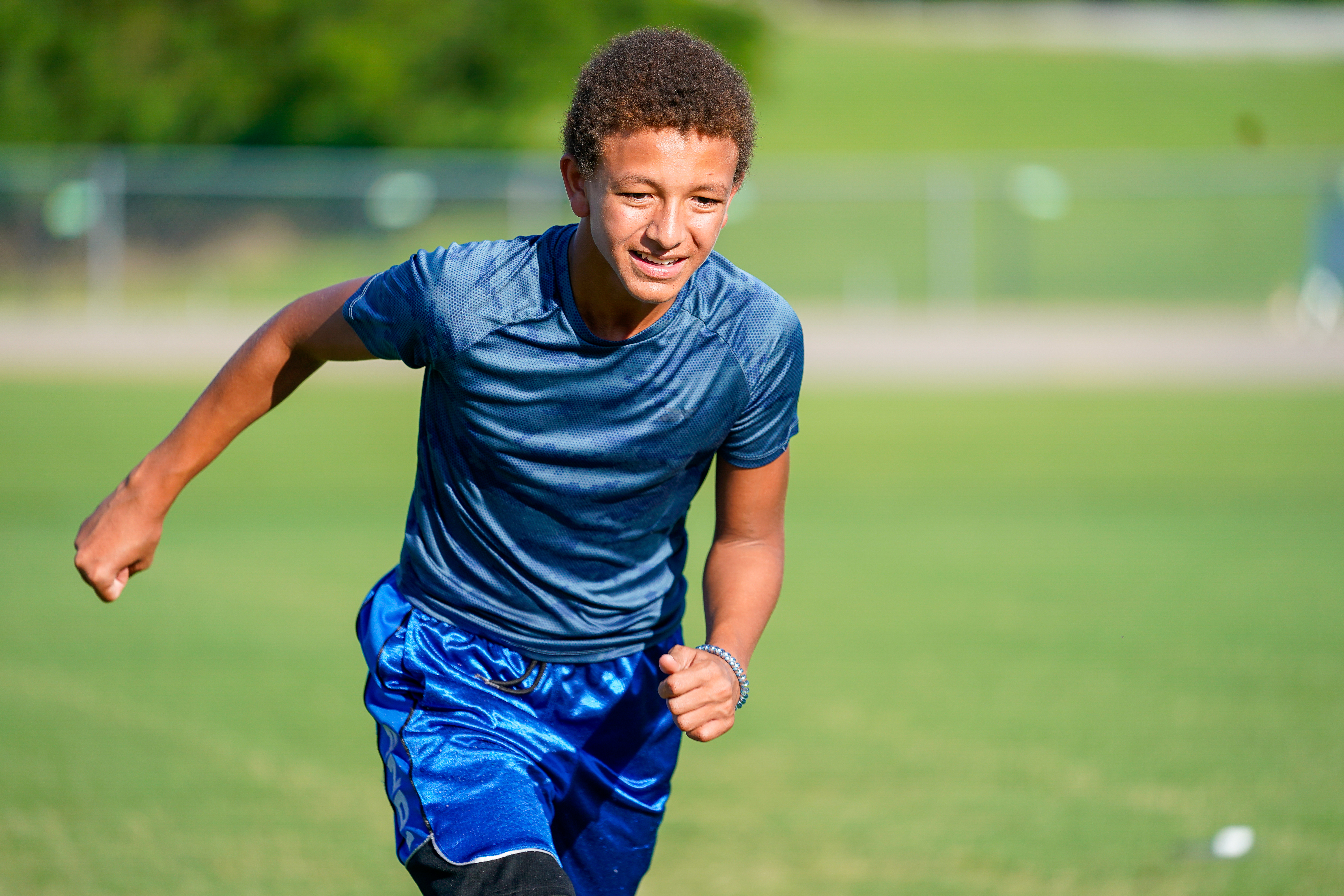 A young boy in a blue shirt and shorts runs across a grassy field with a blurred background of trees and a fence.