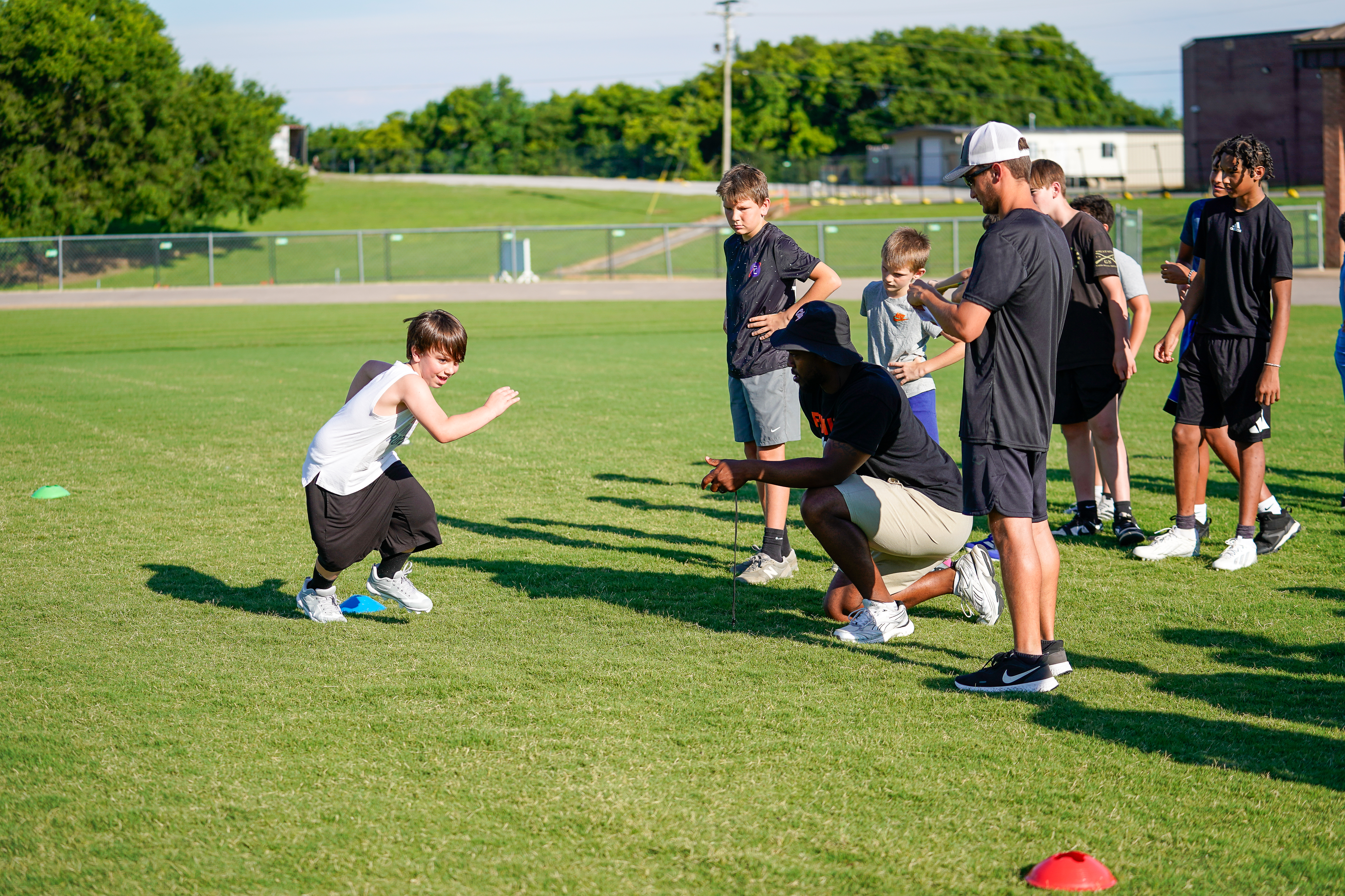 A young boy in a white tank top and black shorts runs on a grassy field while coaches and other boys watch in the background.