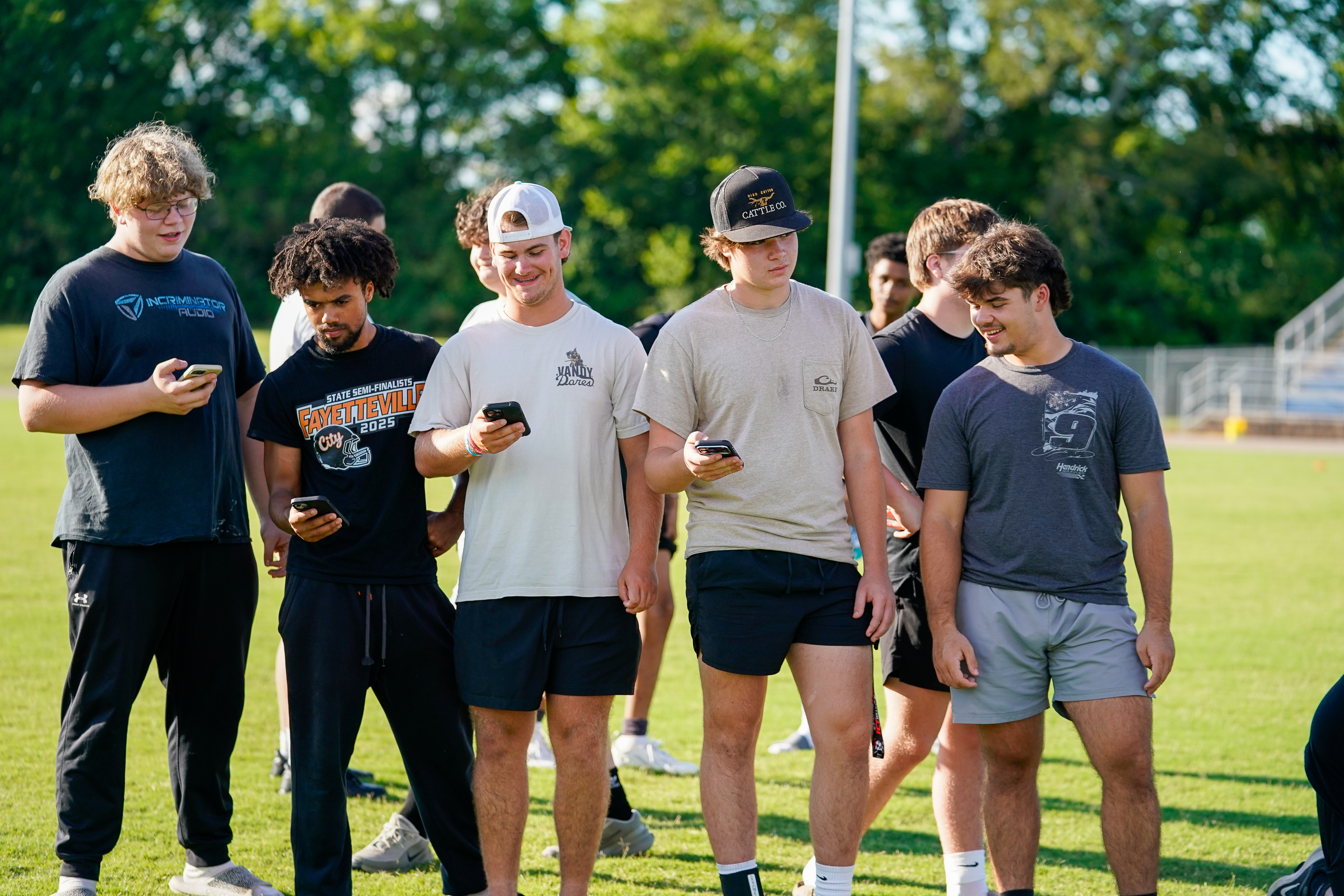 A group of young men in athletic wear stand on a grassy field, looking at their phones, with a blurred stadium and trees in the background.