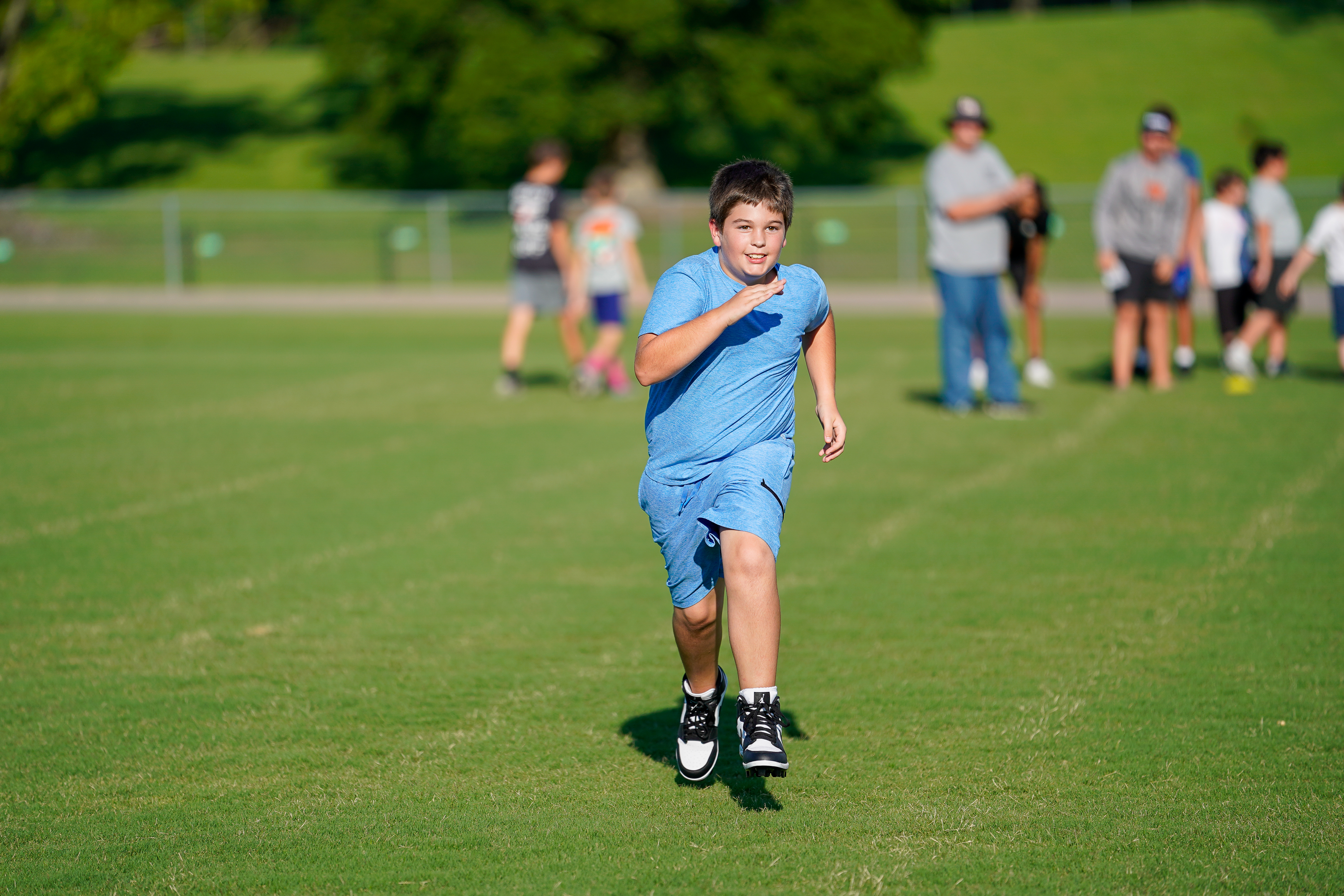 A young boy in a blue shirt and shorts runs on a grassy field with other children and adults in the blurred background.