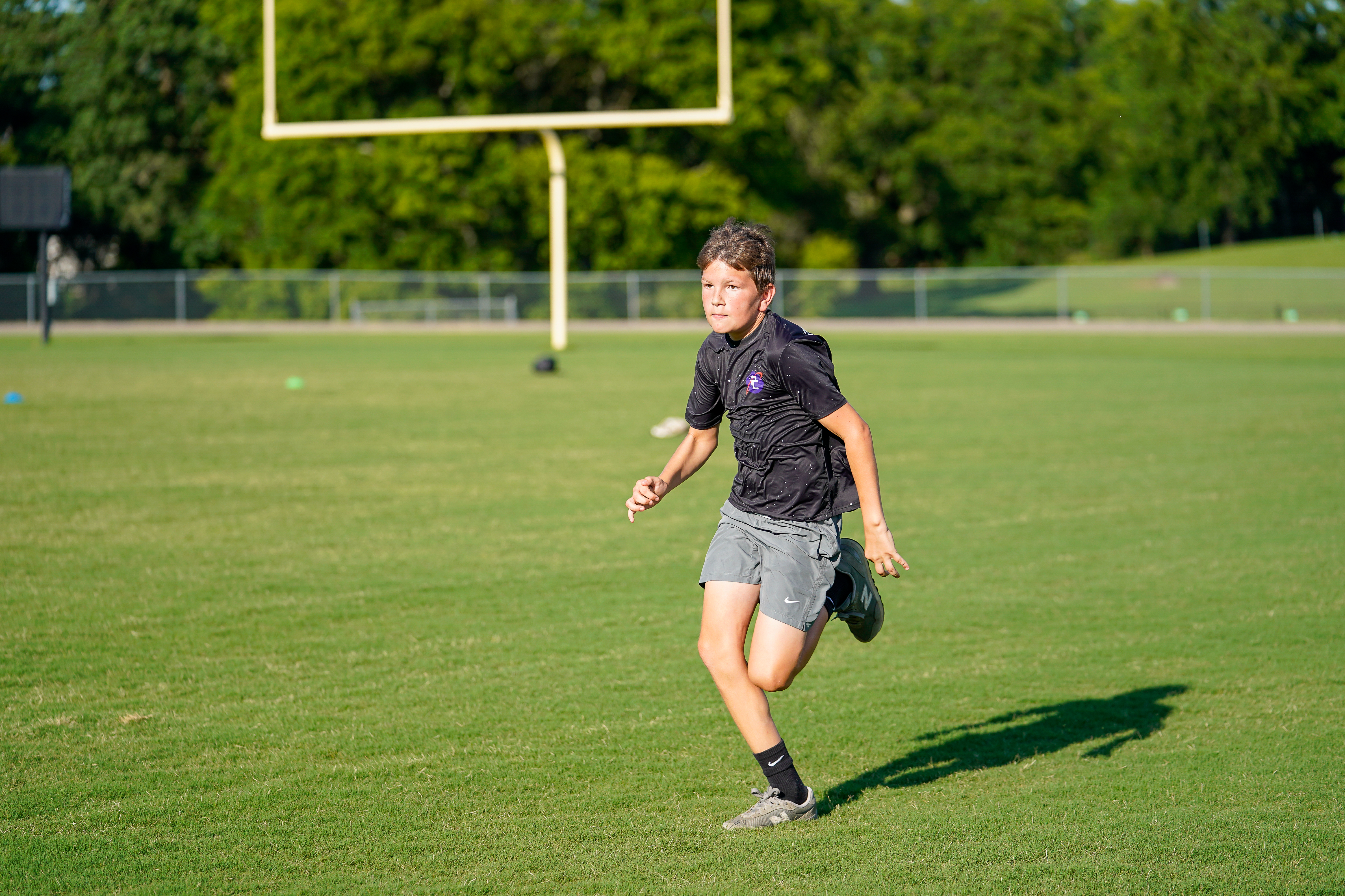 A young boy in athletic wear runs across a green field with a football goalpost and trees in the background.