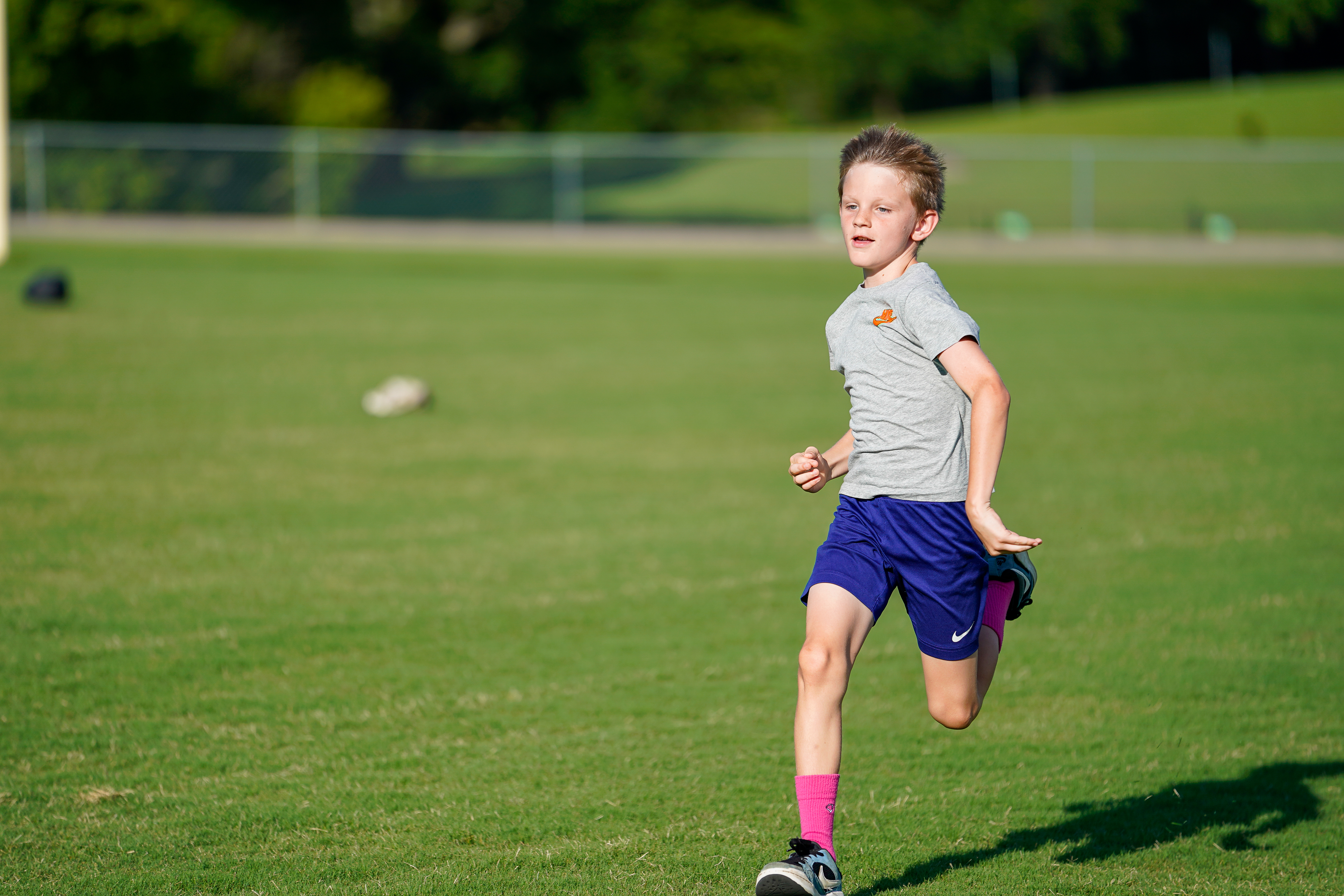 A young boy in a grey shirt and blue shorts runs across a grassy field with a blurred background of trees and a fence.