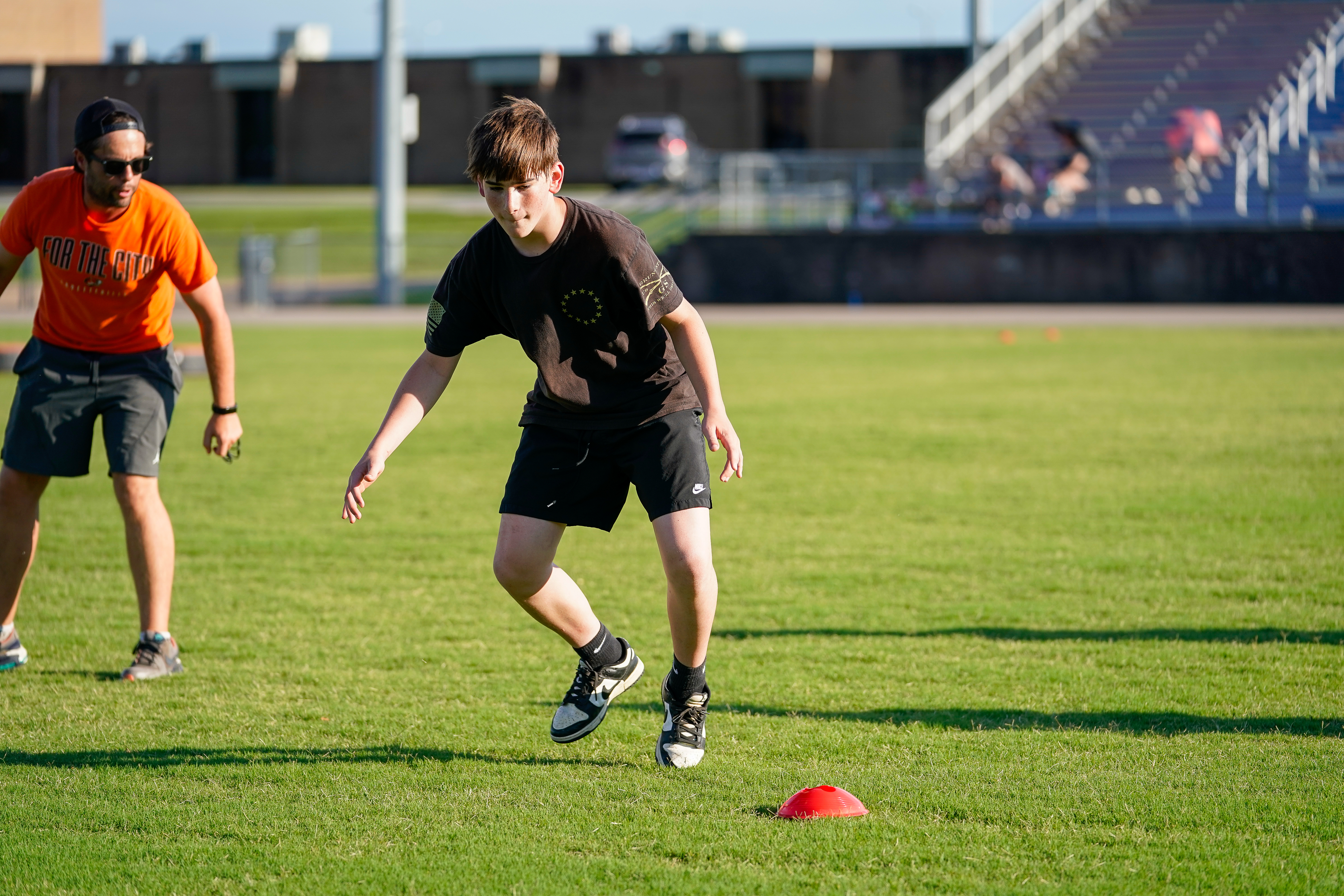 A young athlete in a black shirt and shorts practices drills on a grassy field with a coach in an orange shirt behind him, while stadium bleachers are visible in the background.