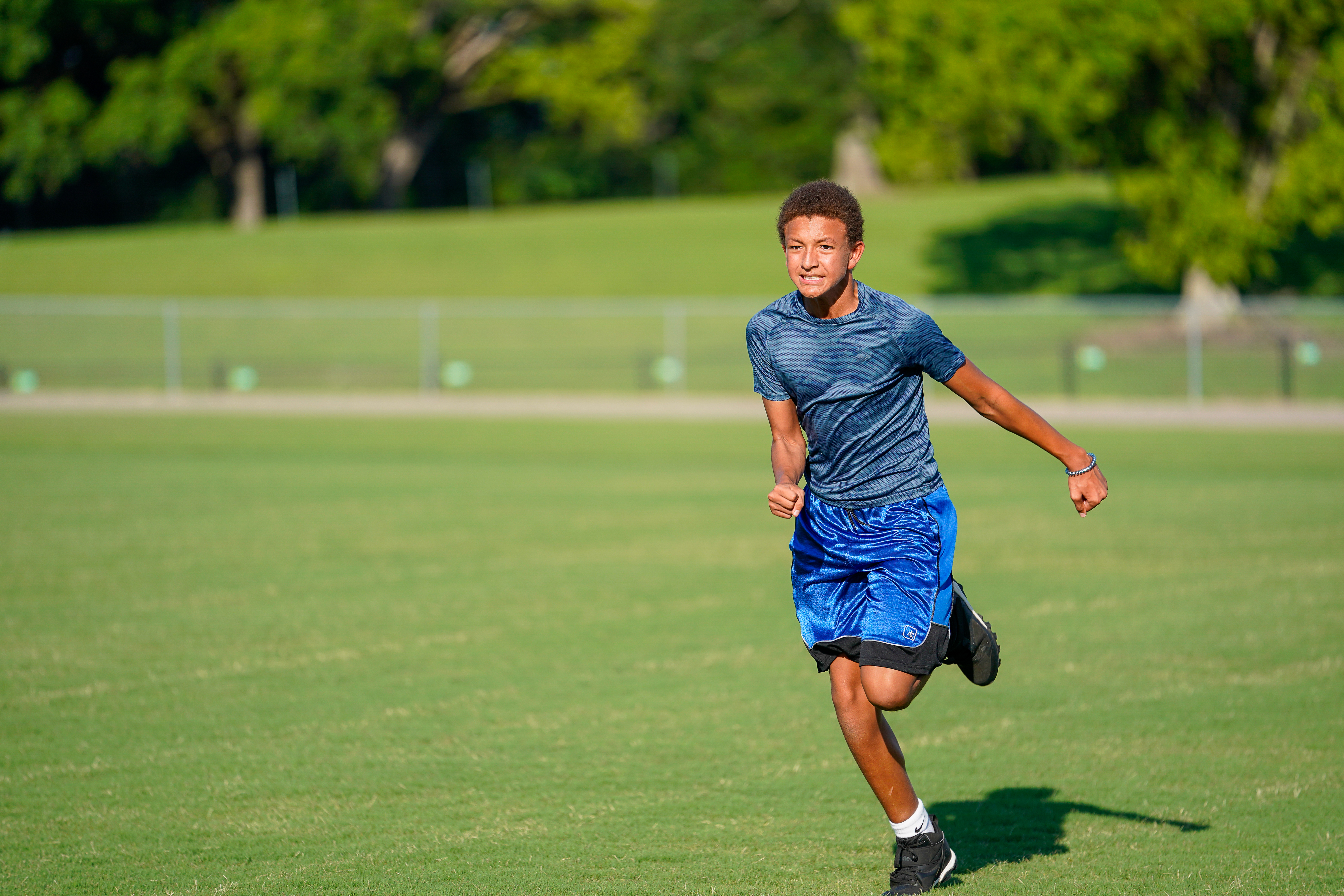 A young man in athletic wear runs across a grassy field with trees and a fence in the background.