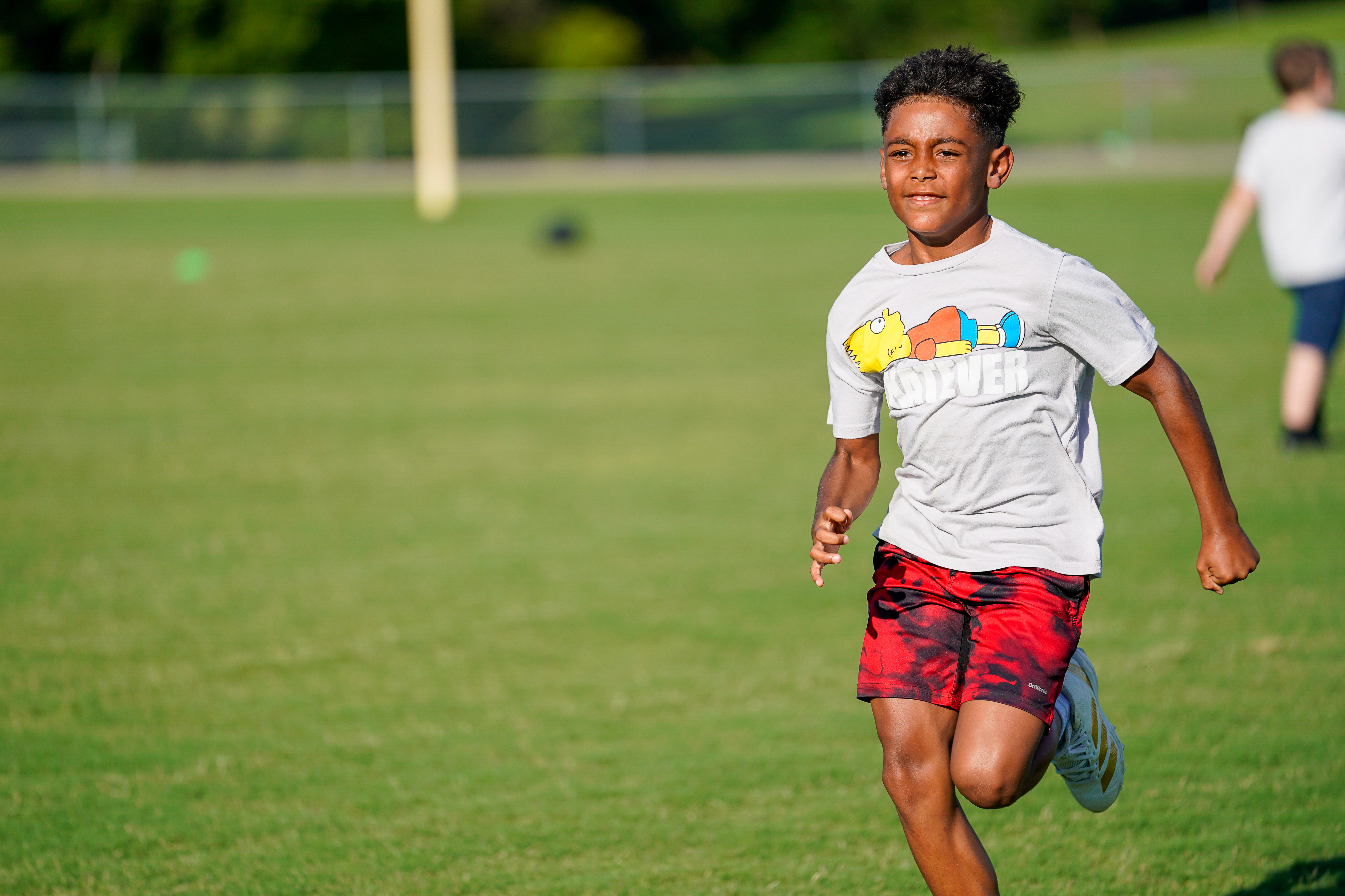 A young boy in a graphic t-shirt and red shorts runs across a grassy field with blurred figures in the background.