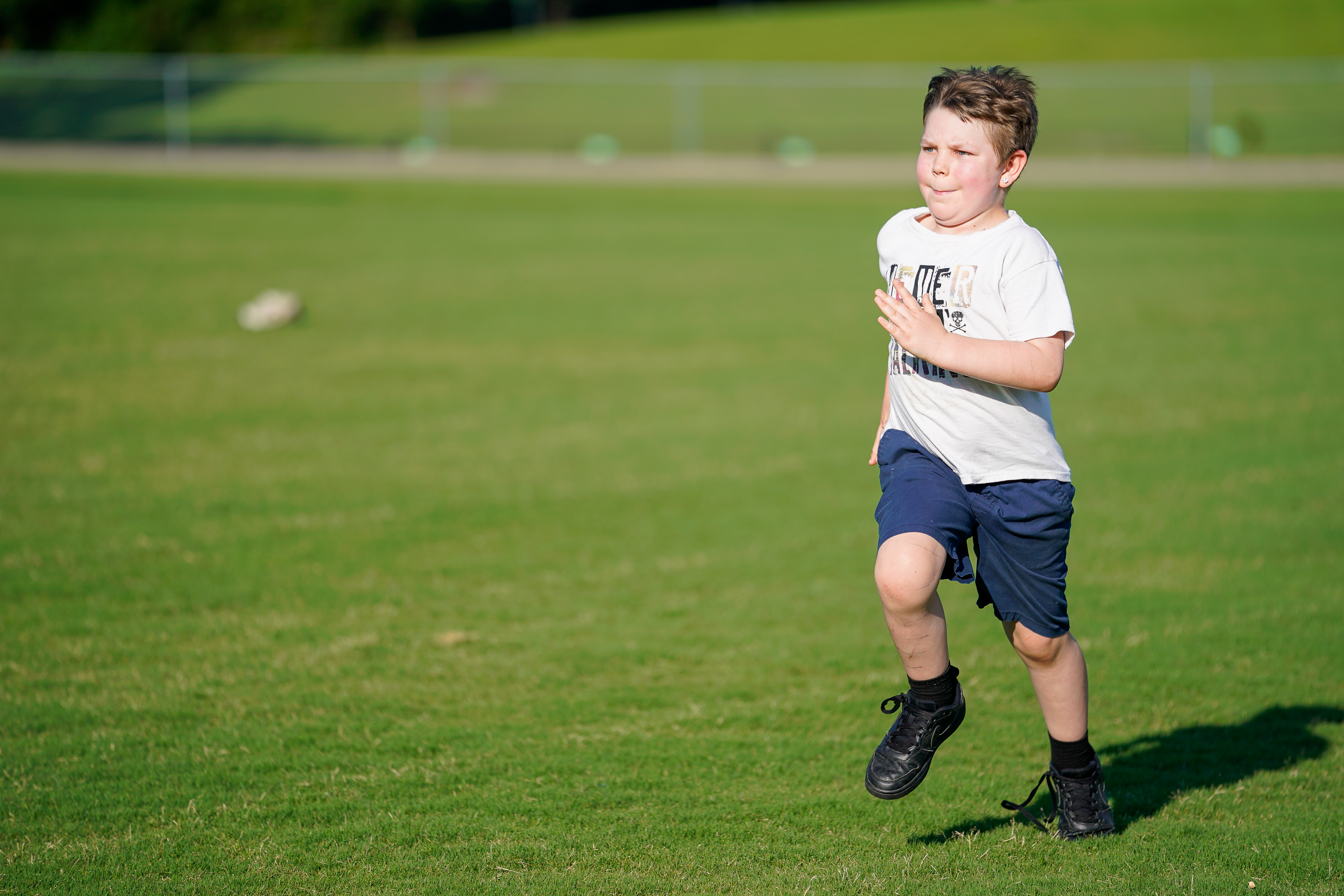 A young boy in a white t-shirt and blue shorts runs across a green grassy field with a blurred background of trees and a fence.