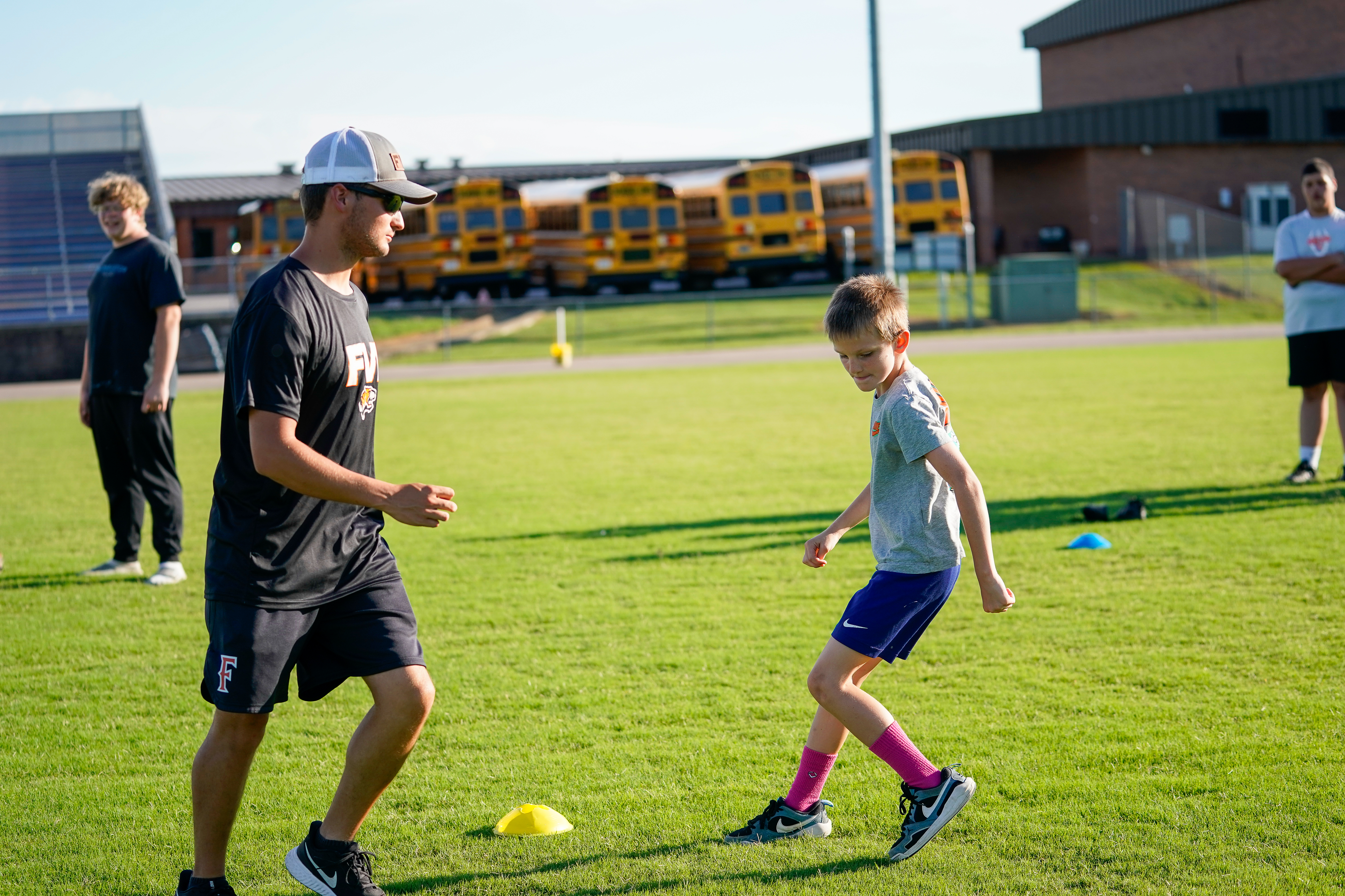 A young boy in a grey shirt and blue shorts practices soccer drills with a coach on a grassy field, with yellow school buses lined up in the background.