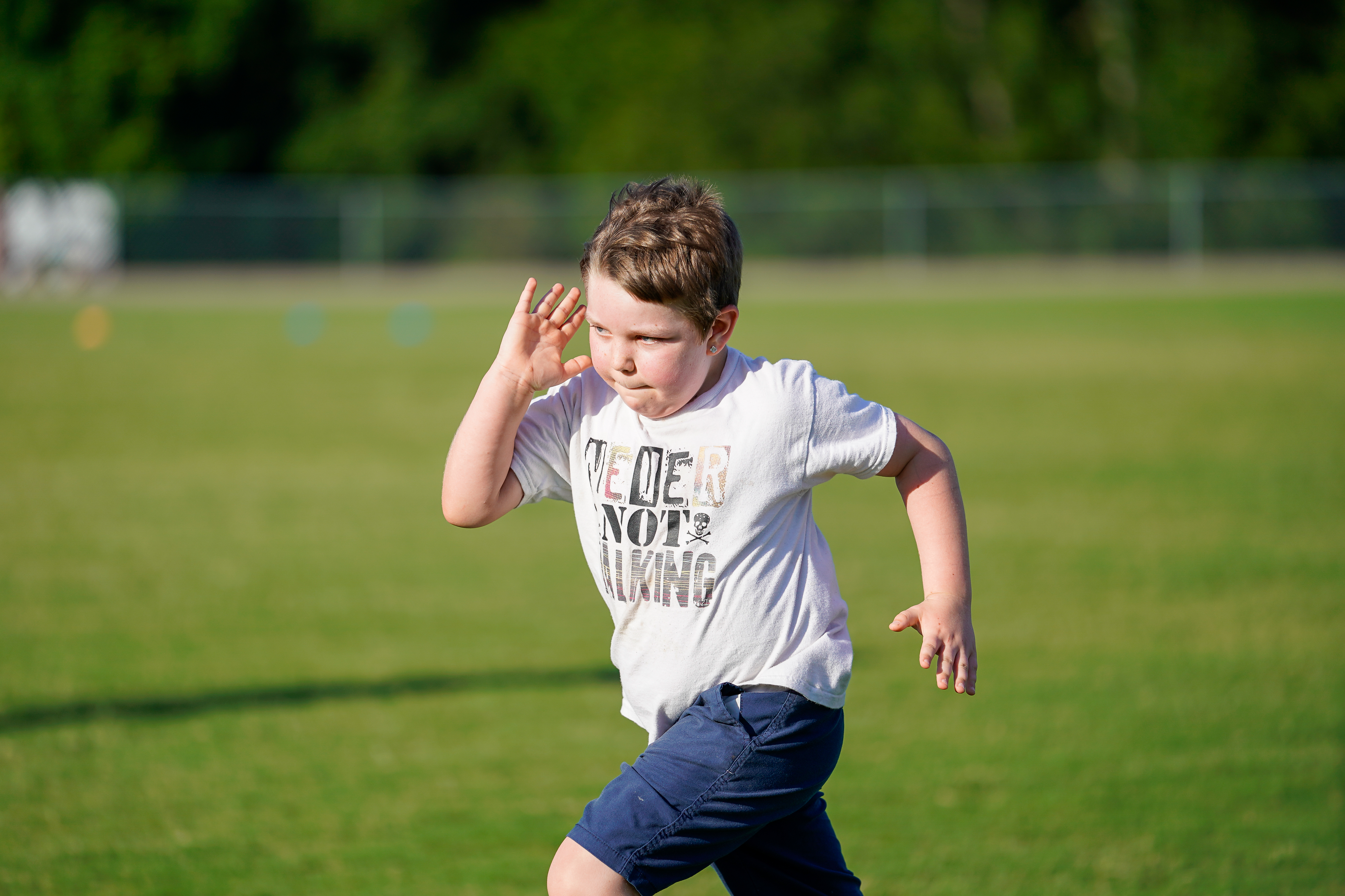 A young boy in a white t-shirt and blue shorts runs across a grassy field with trees in the background.