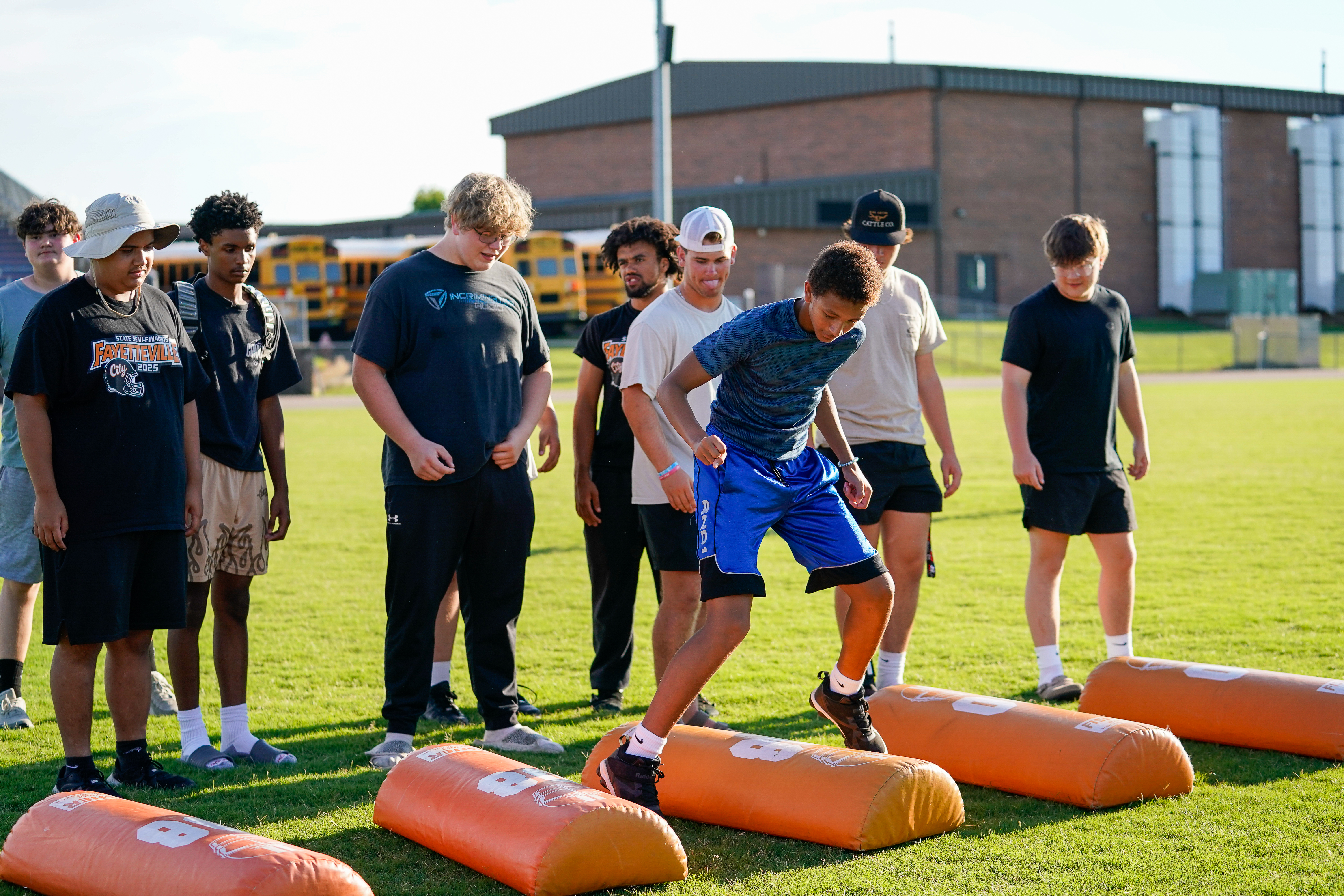 A group of young men in athletic wear practice on a grassy field with orange blocking dummies in the foreground, while yellow school buses and a brick building are visible in the background.