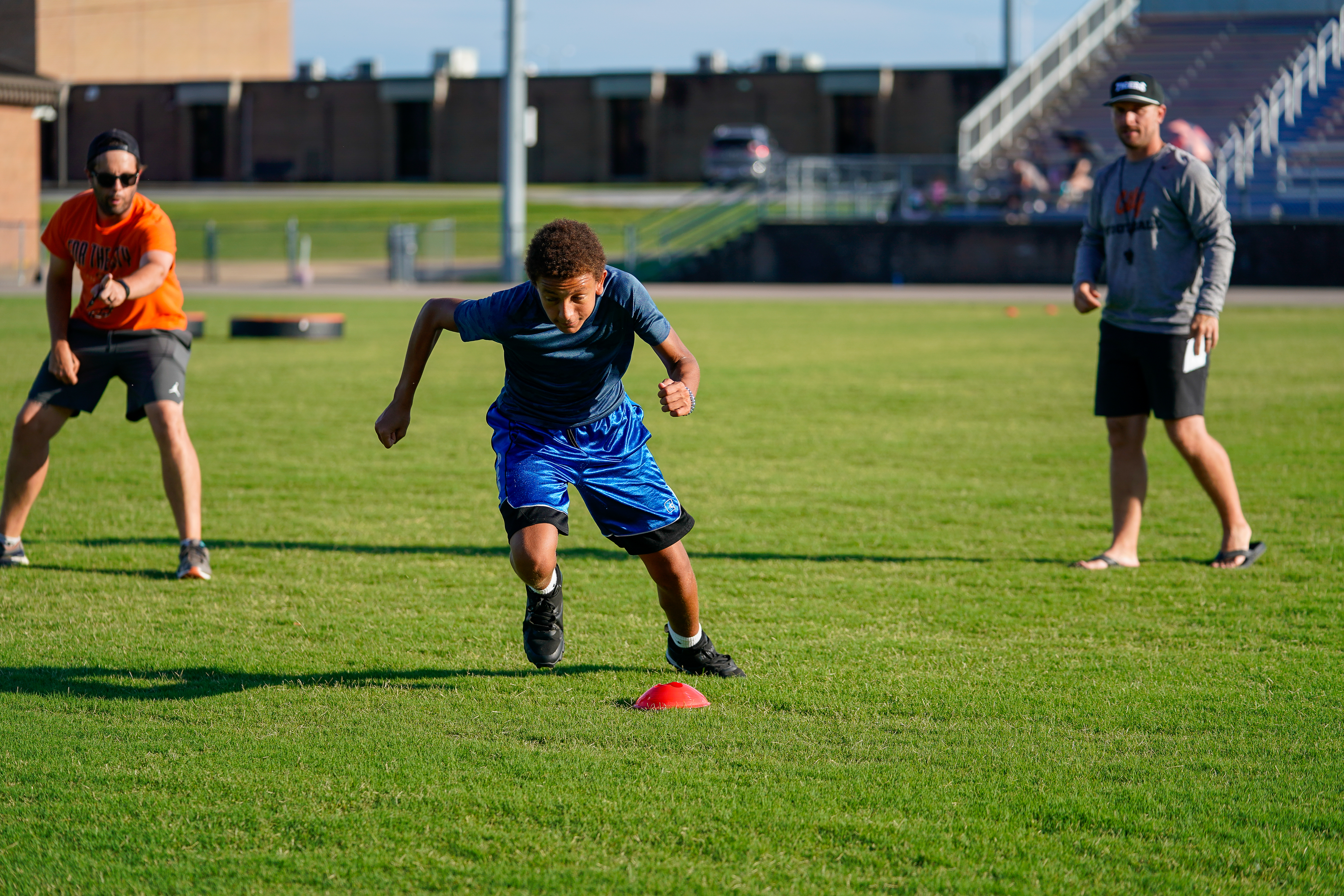 A young boy sprints around a red cone on a grassy field while two adults watch in the background, with bleachers visible beyond them.