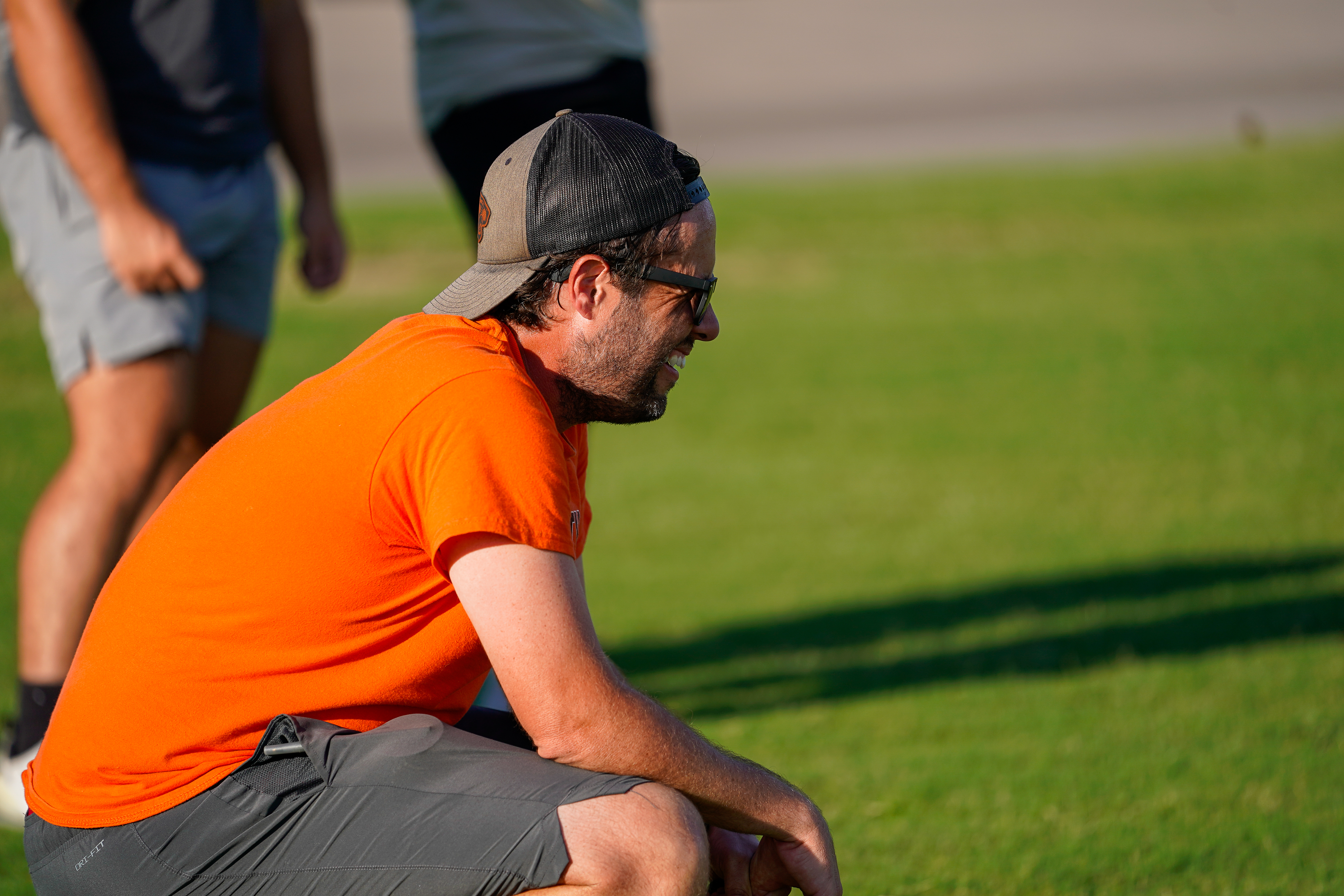 A man in an orange shirt and gray shorts crouches on a grassy field, with other people blurred in the background.
