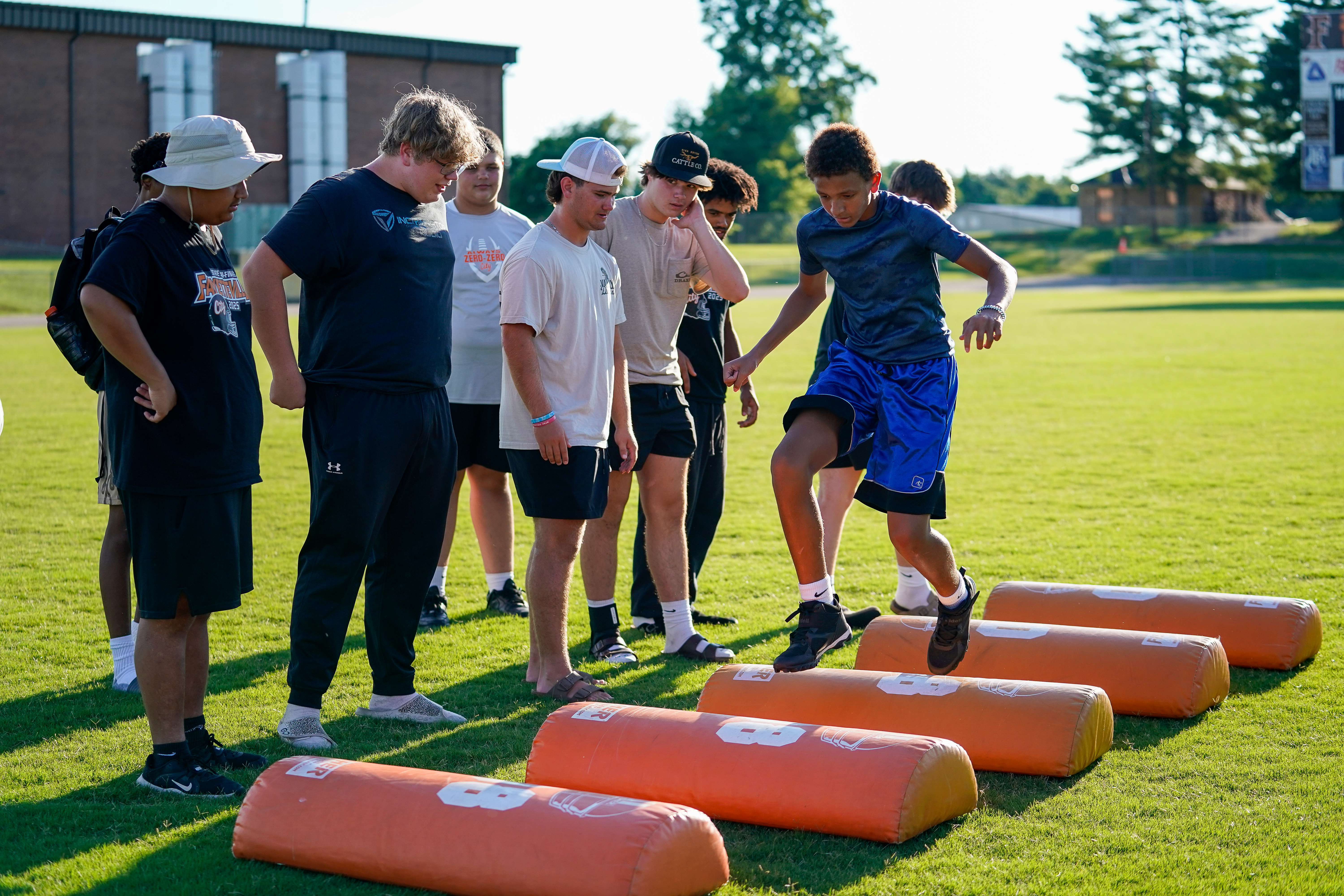 A group of young athletes practice drills over orange hurdles on a grassy field with a building and trees in the background.