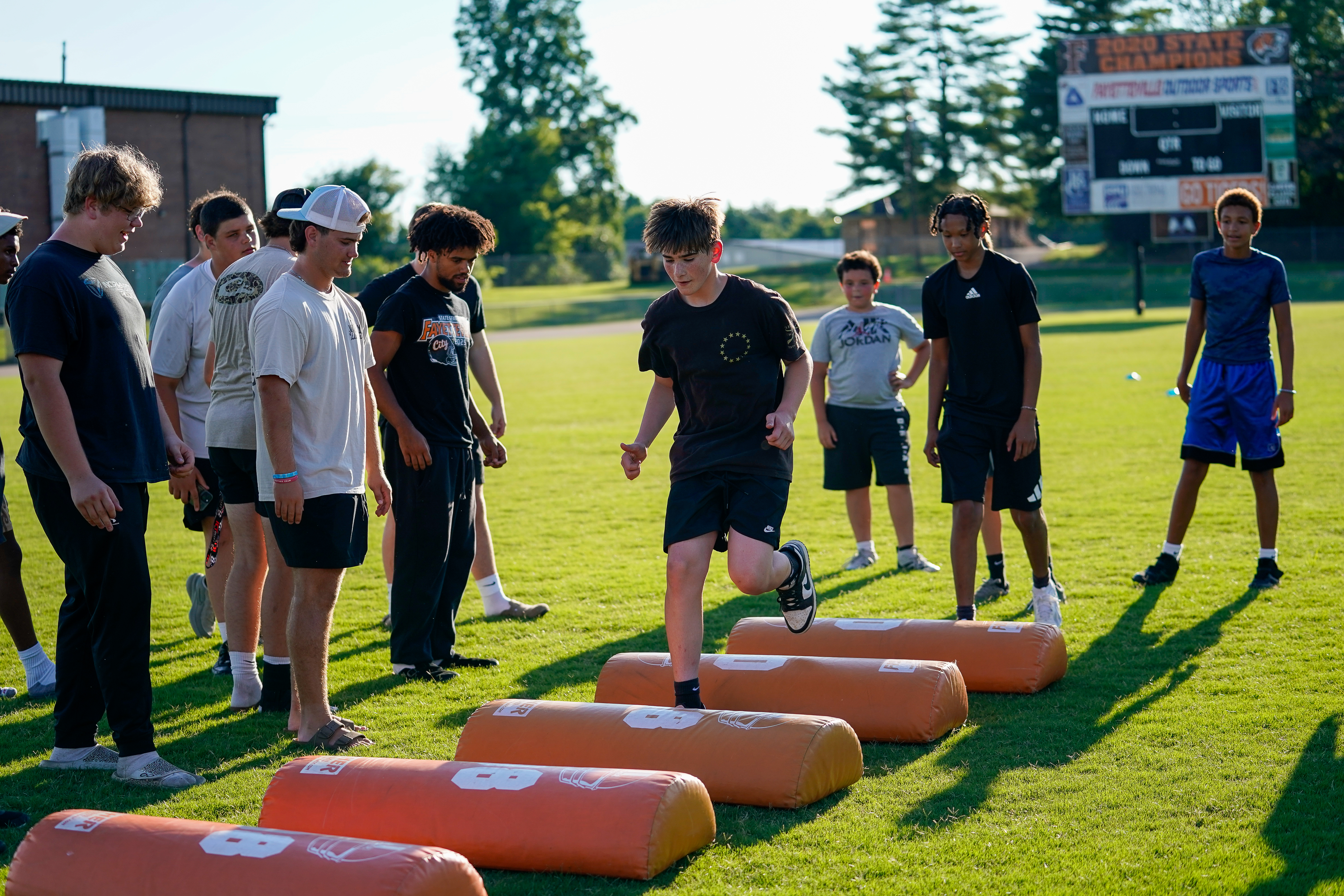 A group of young athletes practice on a grassy field with orange hurdles in the foreground and a scoreboard in the background.