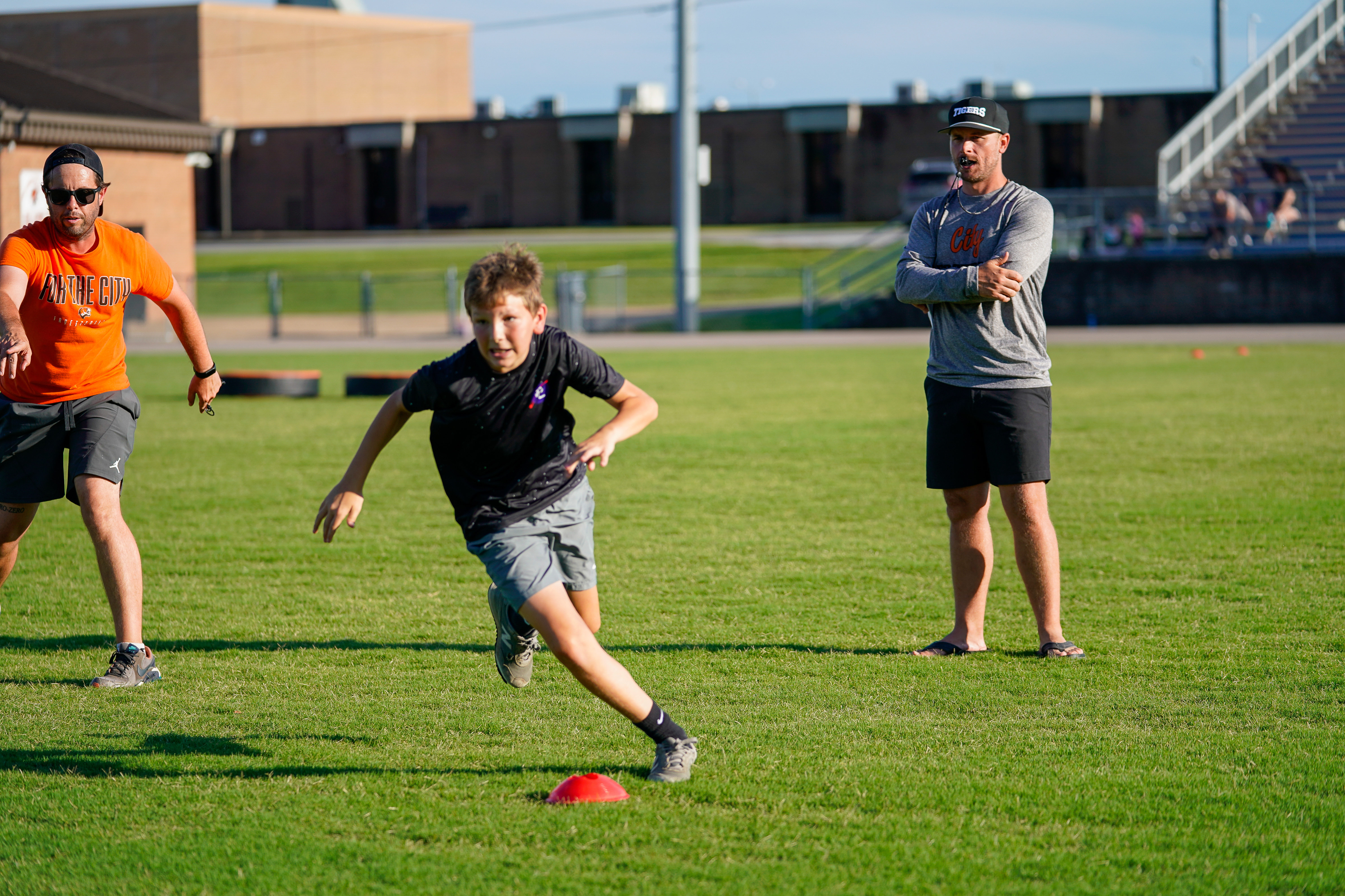 A young boy sprints past a red cone on a grassy field while two men watch in the background, with bleachers visible behind them.