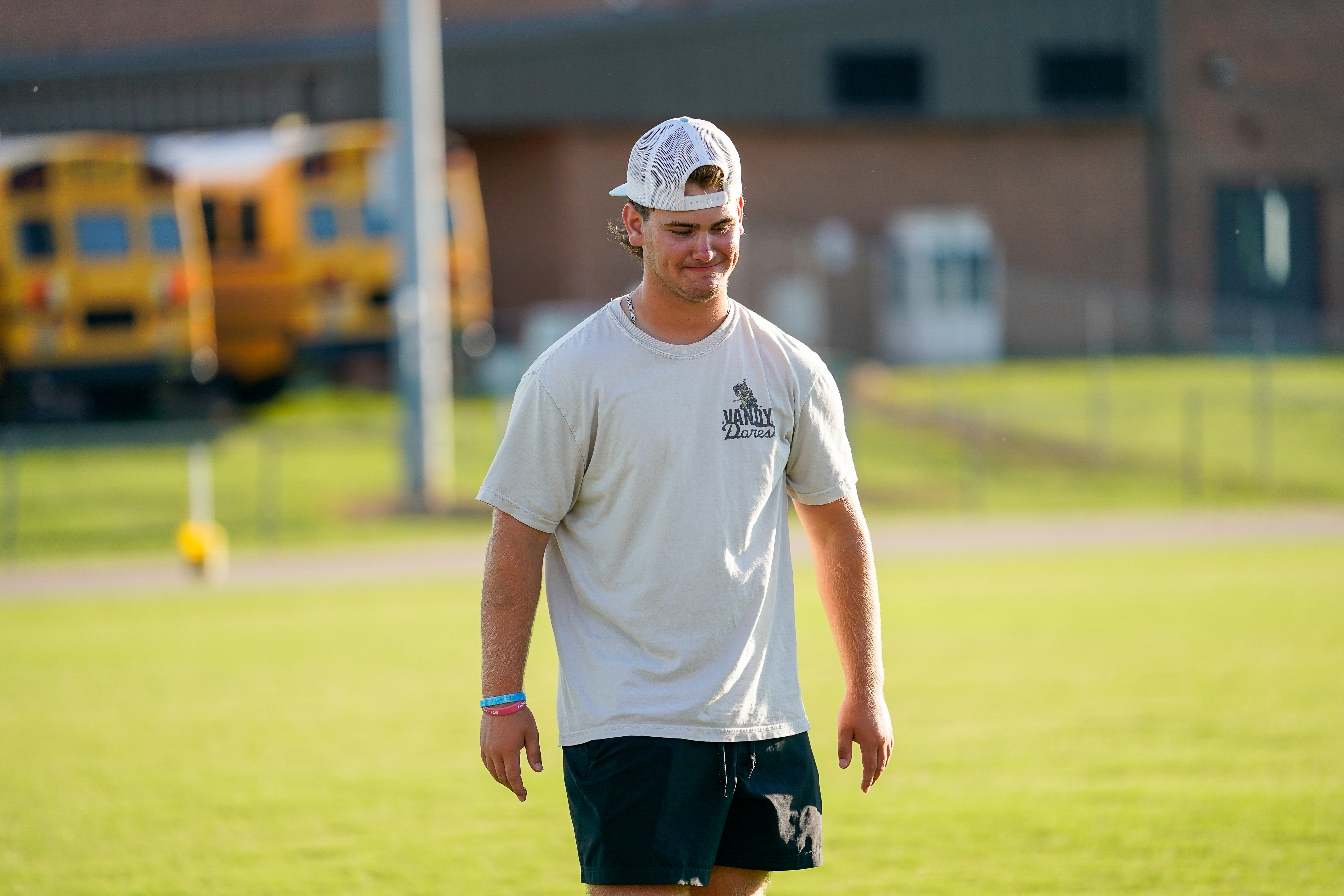 A young man in a white hat and t-shirt stands on a grassy field with a yellow school bus in the blurred background.