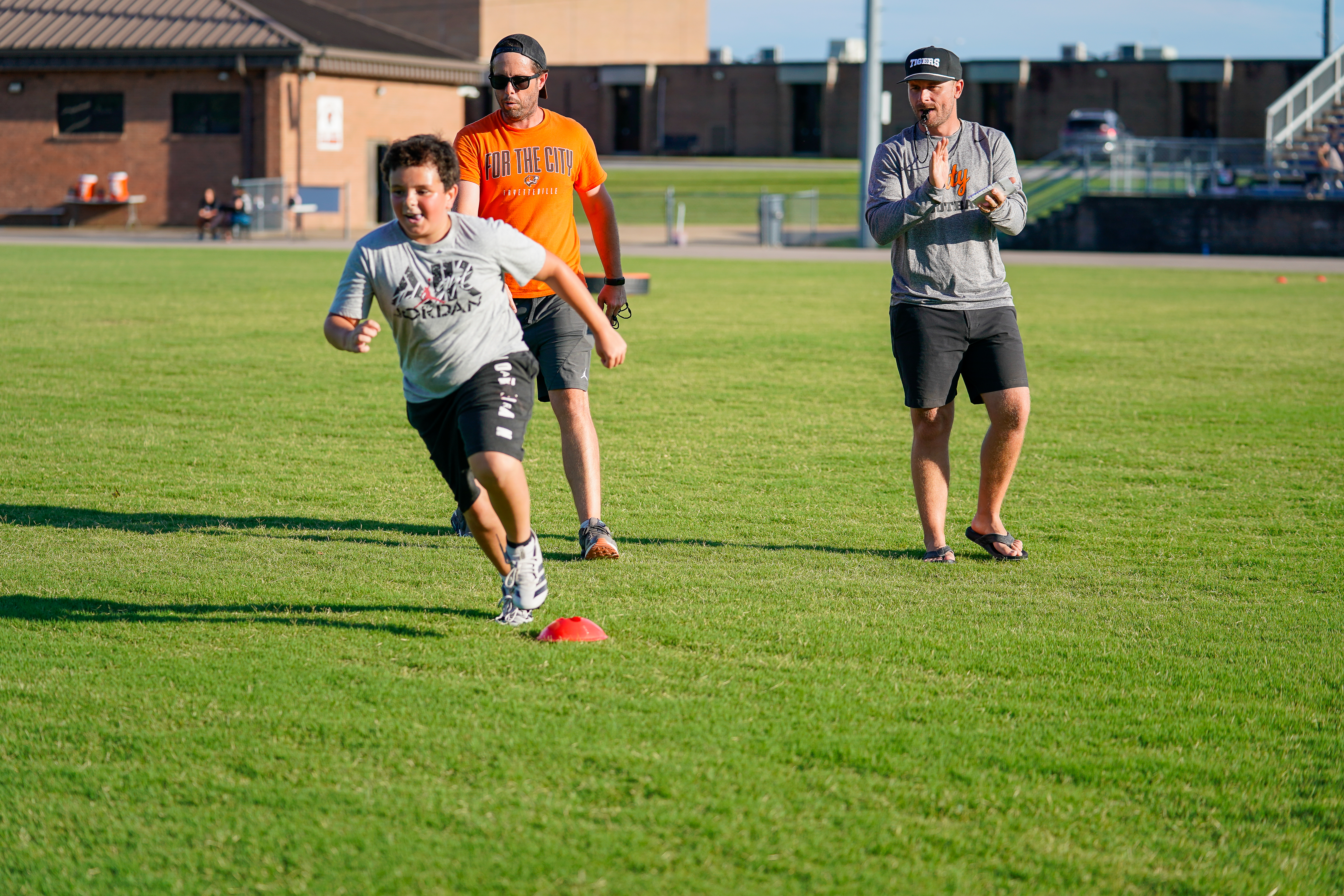 A young boy runs on a grassy field past a red cone, with two men watching him in the background.