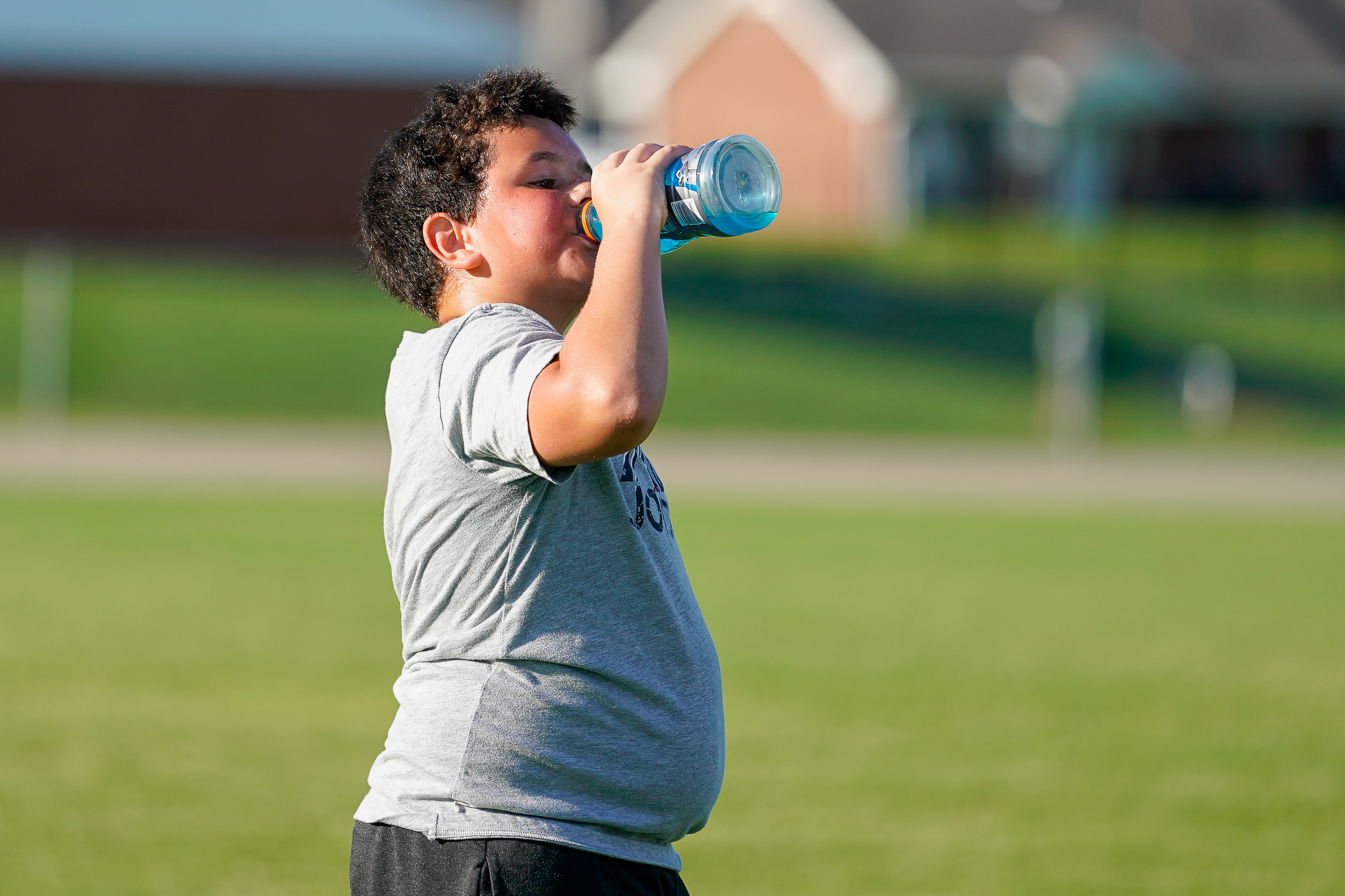 A young boy in a gray t-shirt drinks from a blue water bottle on a grassy field with a building in the blurred background.