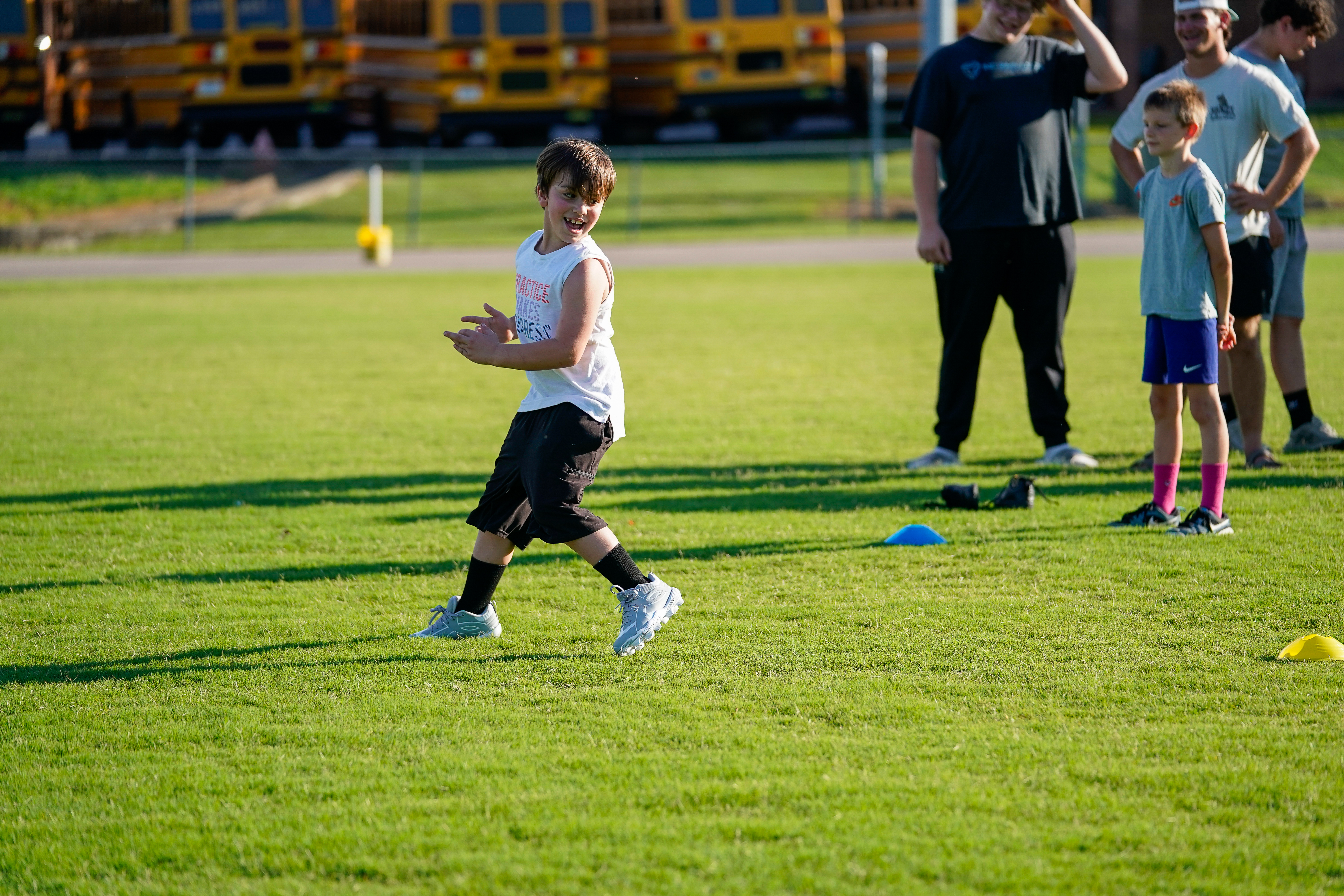 A young boy in the foreground runs on a grassy field with several people and yellow school buses in the background.